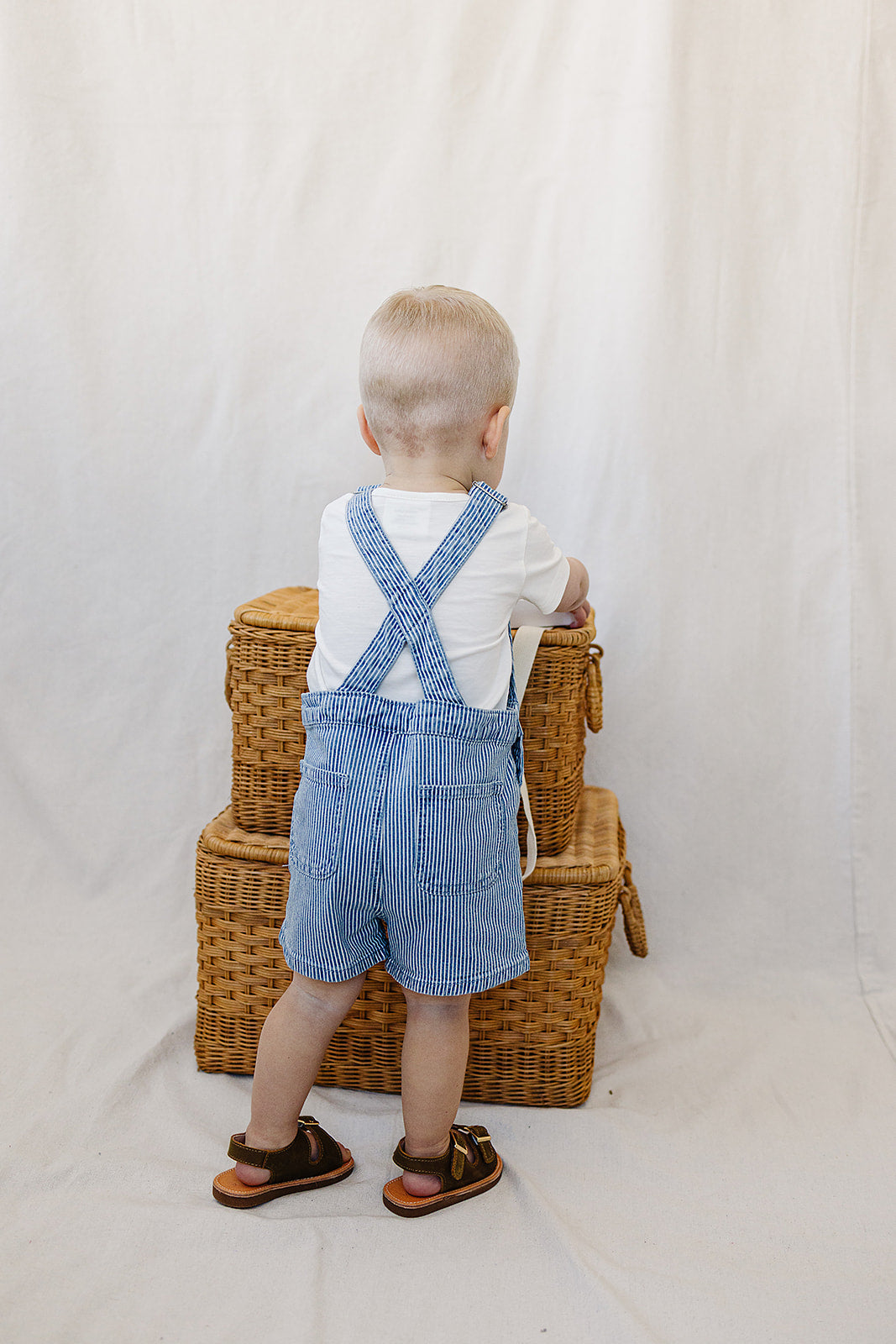 Toddler in blue stripe short overalls with white t-shirt, standing by wicker baskets, perfect summer outfit.