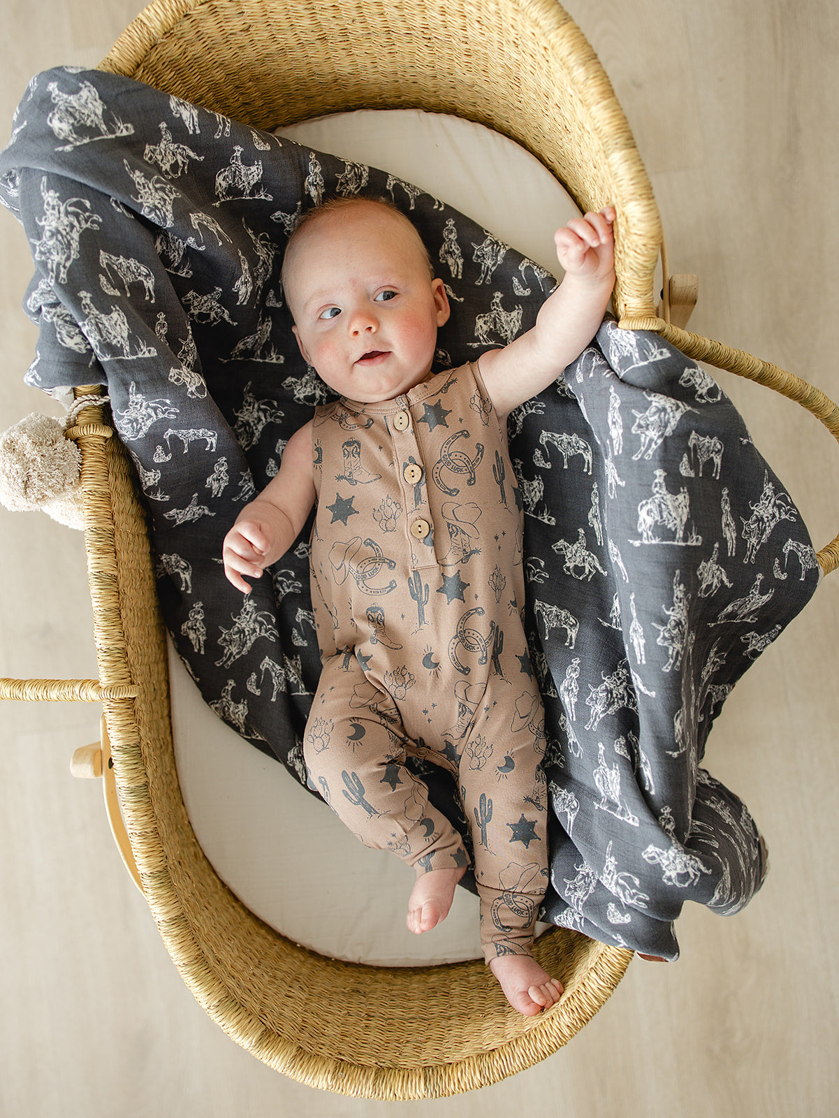 Baby wearing a Wild West Tank Romper with cowboy boot print, lying on a gray blanket in a cozy bassinet.