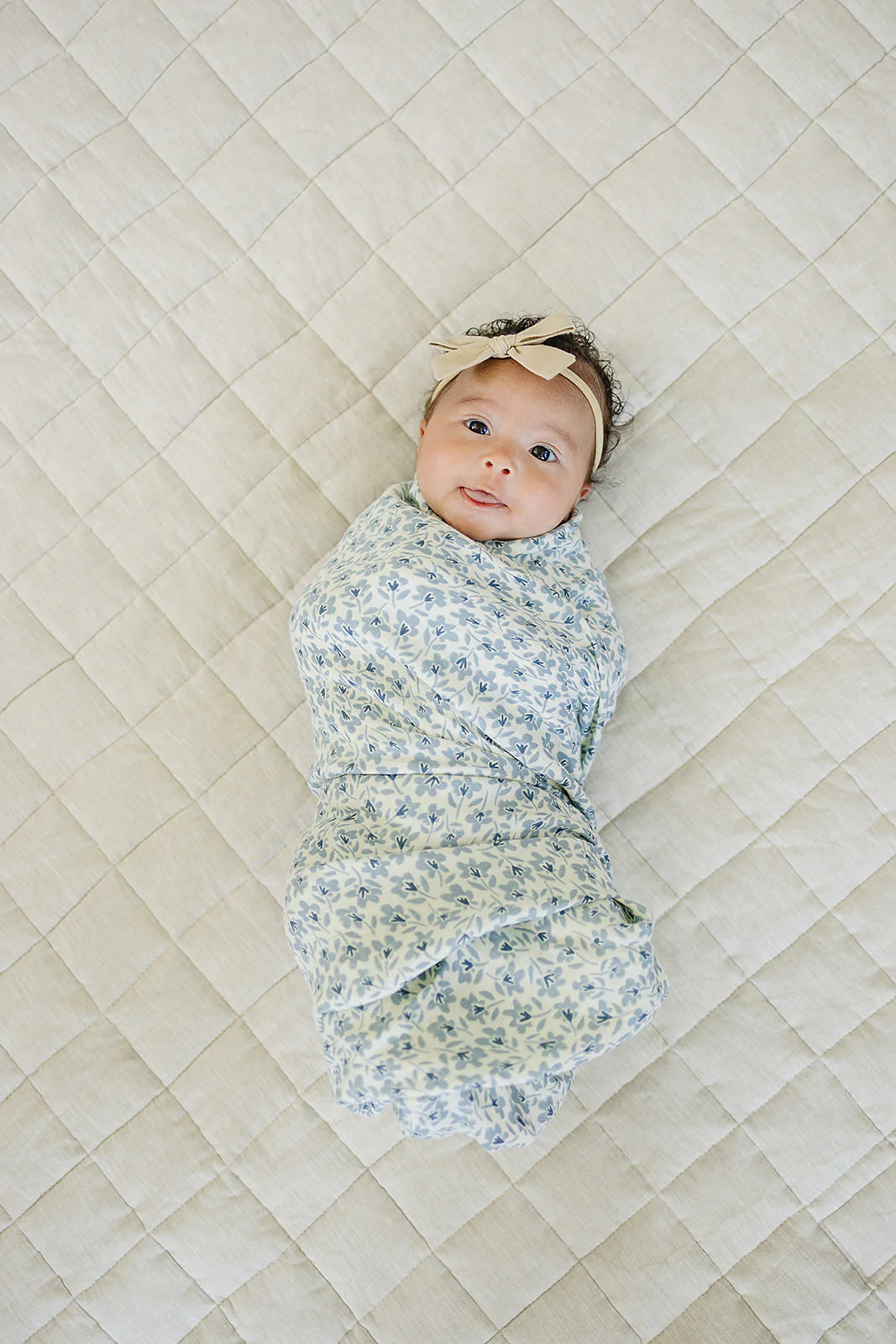 Baby wrapped in a blue blossom muslin swaddle blanket on a quilted surface.