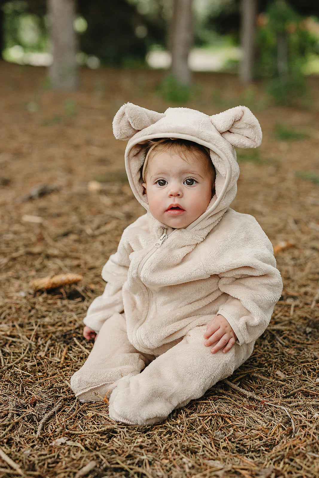 Baby wearing a baby fleece bear hooded romper with ears, sitting on forest ground, looking adorable and cozy.