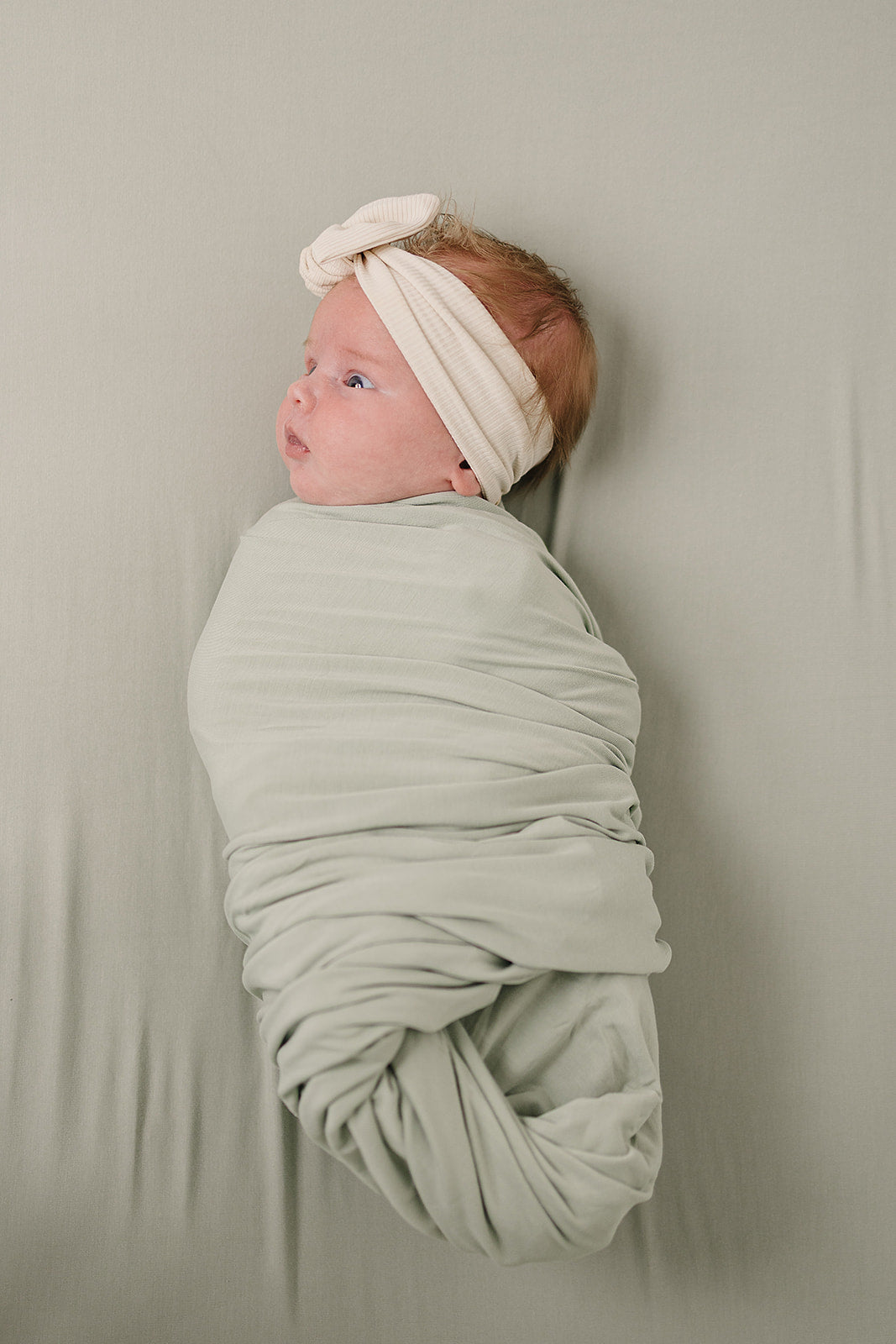 Baby wrapped in the Bamboo Stretch Swaddle Sage with a headband on a soft surface.