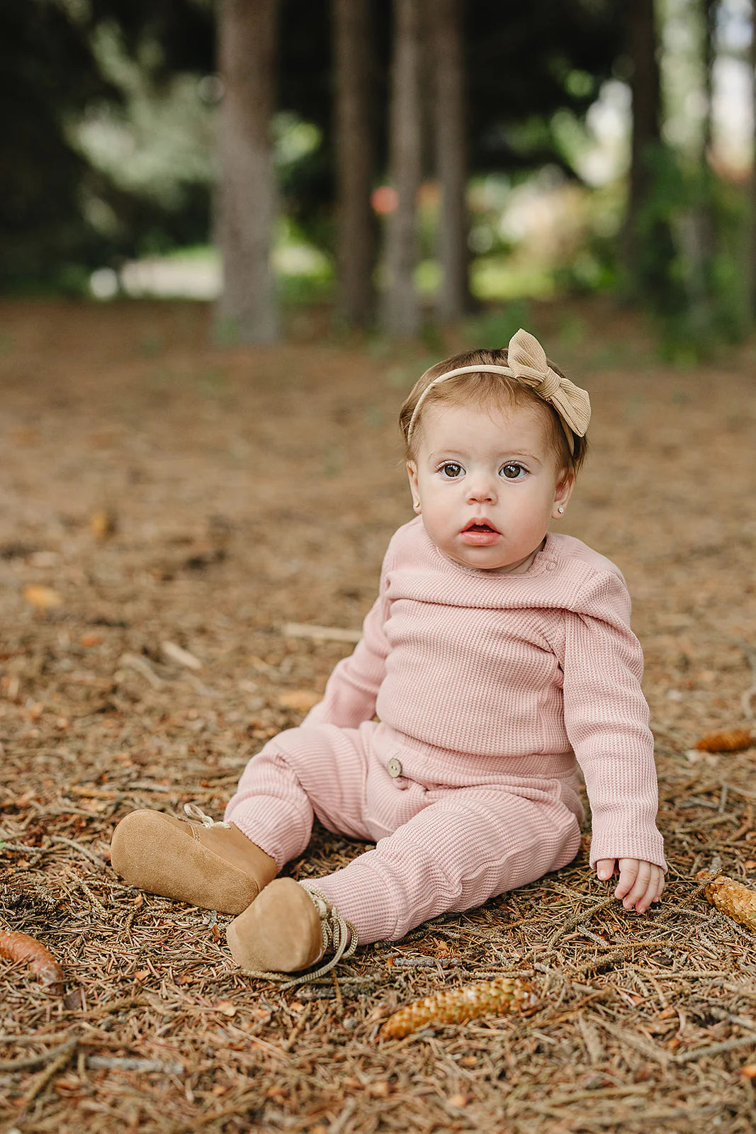 Baby wearing a pink waffle knit baby bodysuit set in a natural outdoor setting, surrounded by pine leaves.