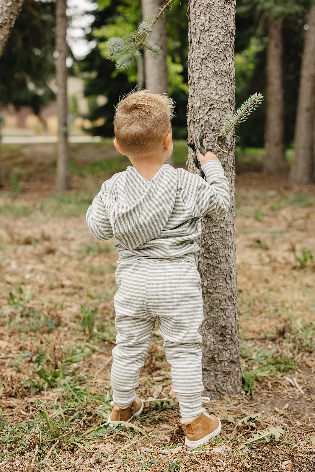 Child wearing grey striped baby and toddler lounge set with hoodie in outdoor setting, playing near a tree.