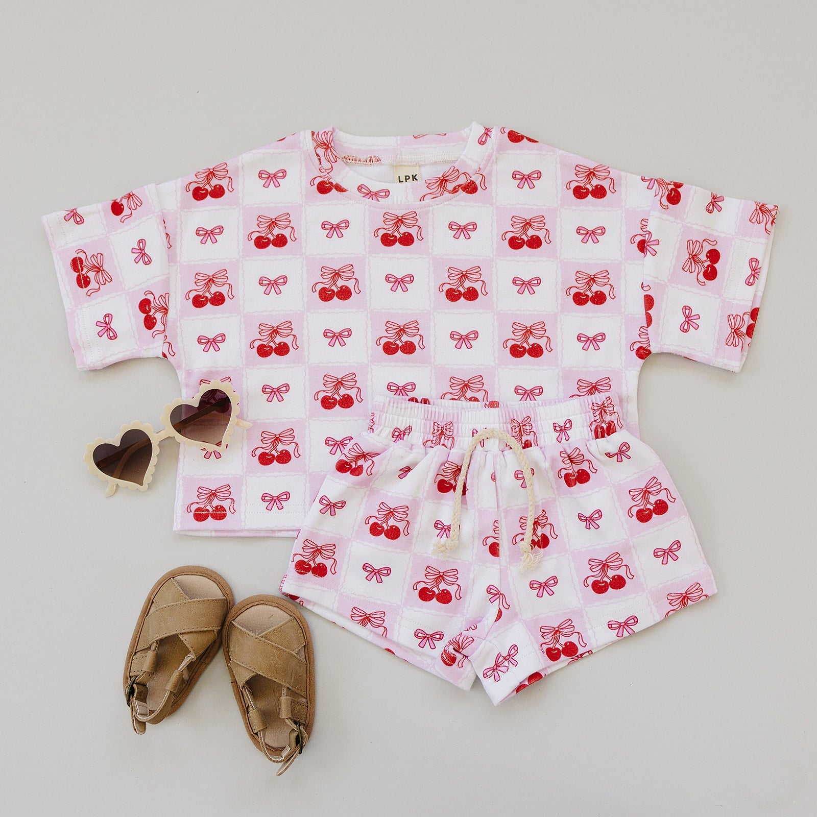 Checkered Cherry shorts set with cherry print and matching top, styled with sandals and heart-shaped sunglasses.