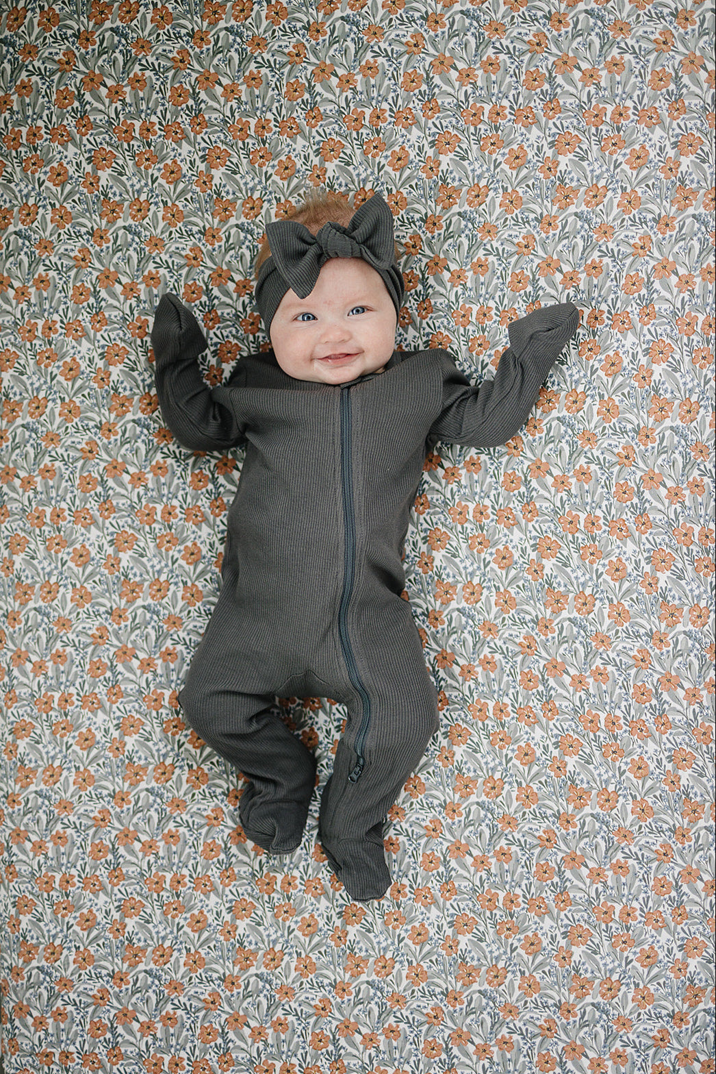 Baby lying on a Dainty Meadow Floral Bamboo Crib Sheet, wearing a gray outfit with a bow.