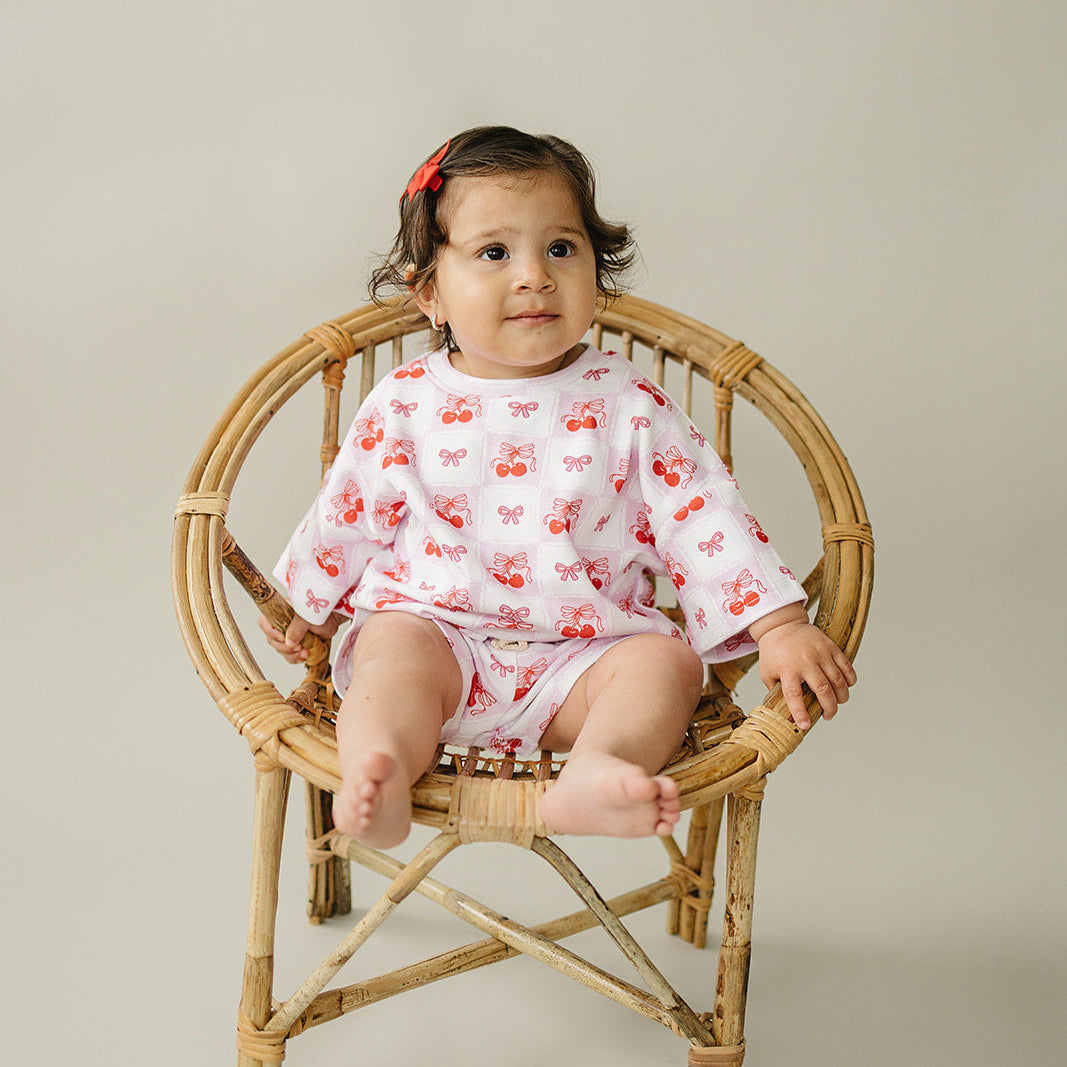 Toddler wearing checkered cherry shorts set, sitting on a rattan chair, showcasing playful cherry motifs.