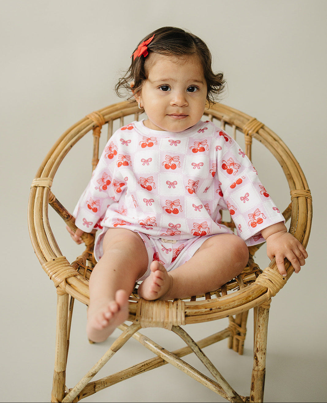 Toddler wearing a checkered cherry shorts set, seated in a rattan chair, showcasing sweet and trendy summer fashion.
