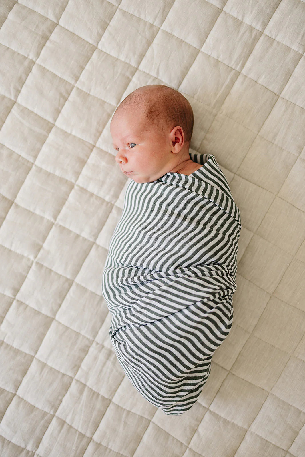 A newborn wrapped in a green stripes muslin swaddle on a soft, quilted blanket.