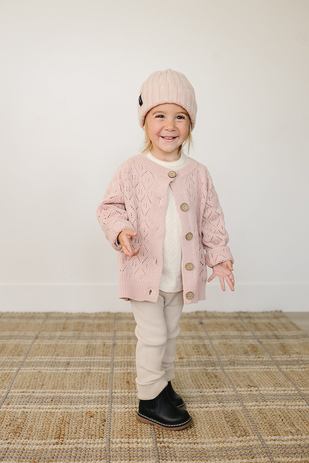 Smiling toddler in a light pink chunky knit baby beanie and matching cardigan, cozy winter outfit.