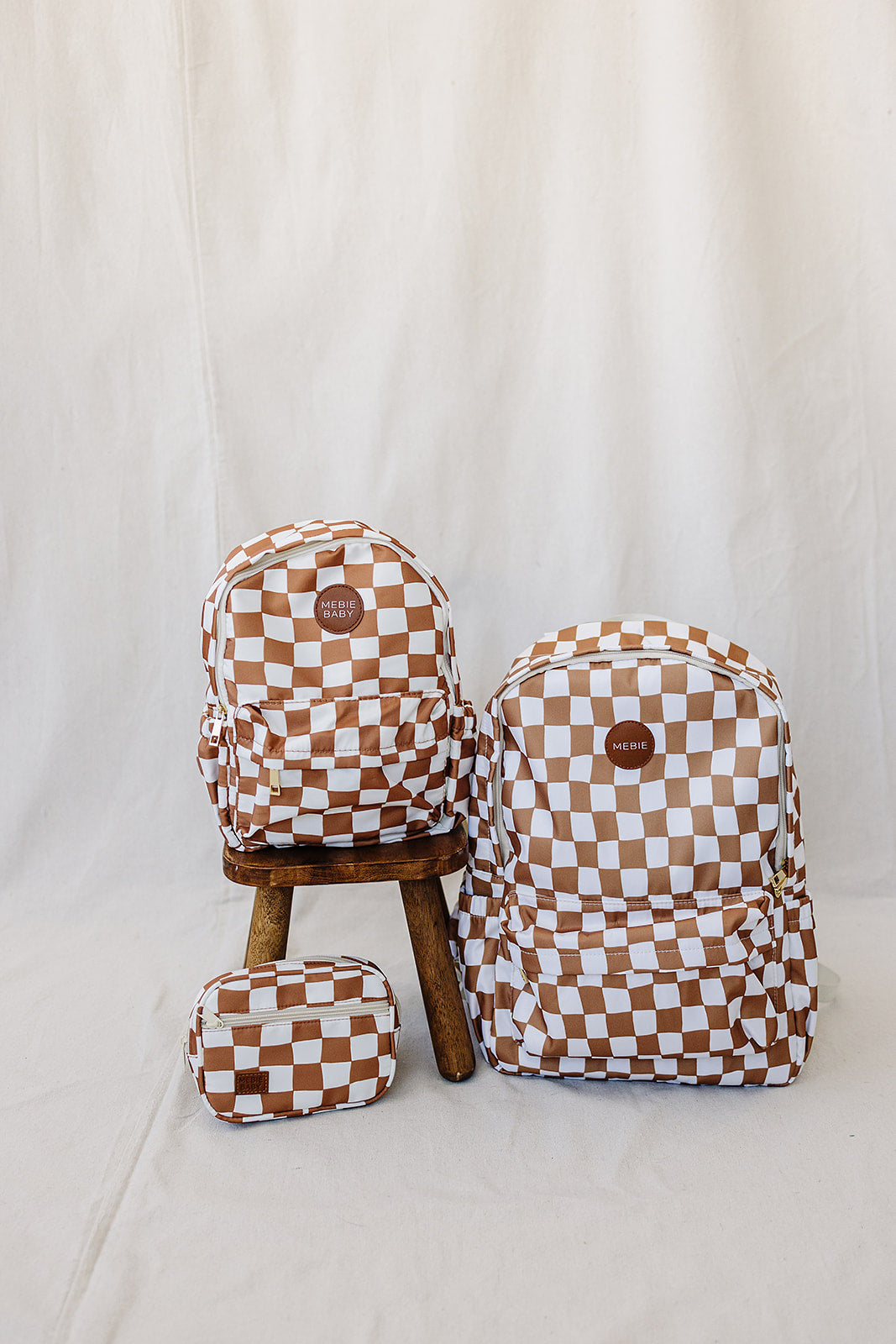 Rust checkered mini fanny pack and two matching backpacks on a wooden stool against a neutral background.