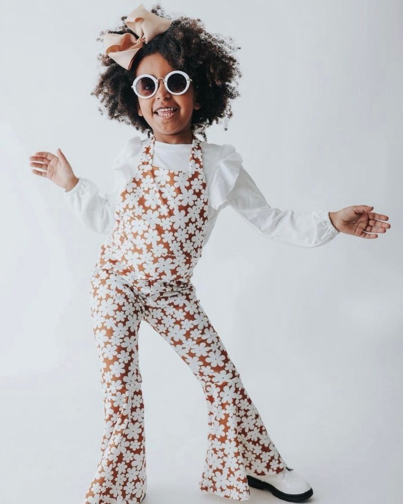 Girl wearing Andi pleated bell bottom jumpsuit in floral print, styled with sunglasses and rosy bow, showcasing retro fashion.