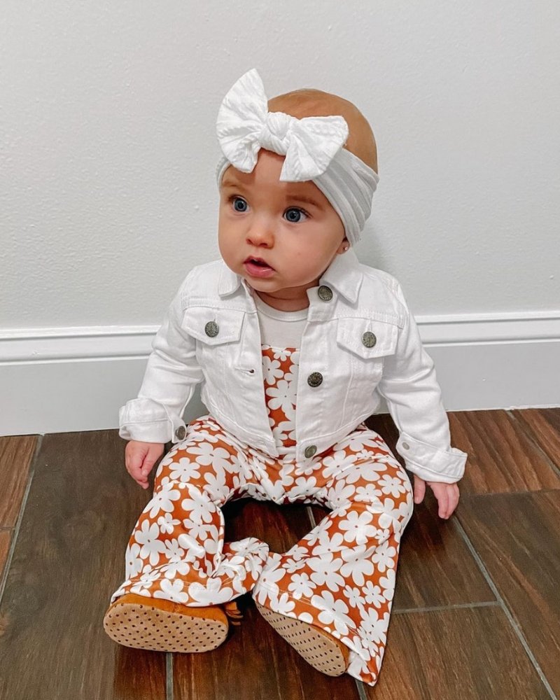 Baby girl in Andi pleated bell bottom jumpsuit with floral print and white denim jacket, accessorized with a bow headband.
