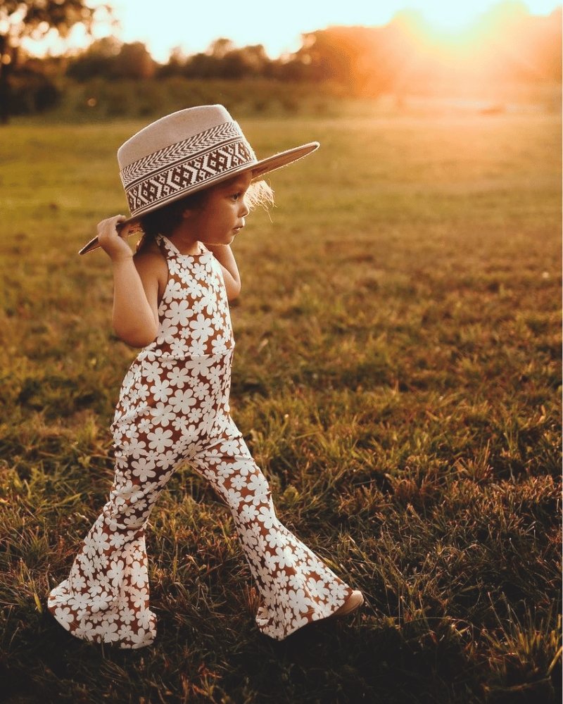 Little girl wearing Andi pleated bell bottom jumpsuit in floral print with a sun hat, enjoying a sunny outdoor setting.