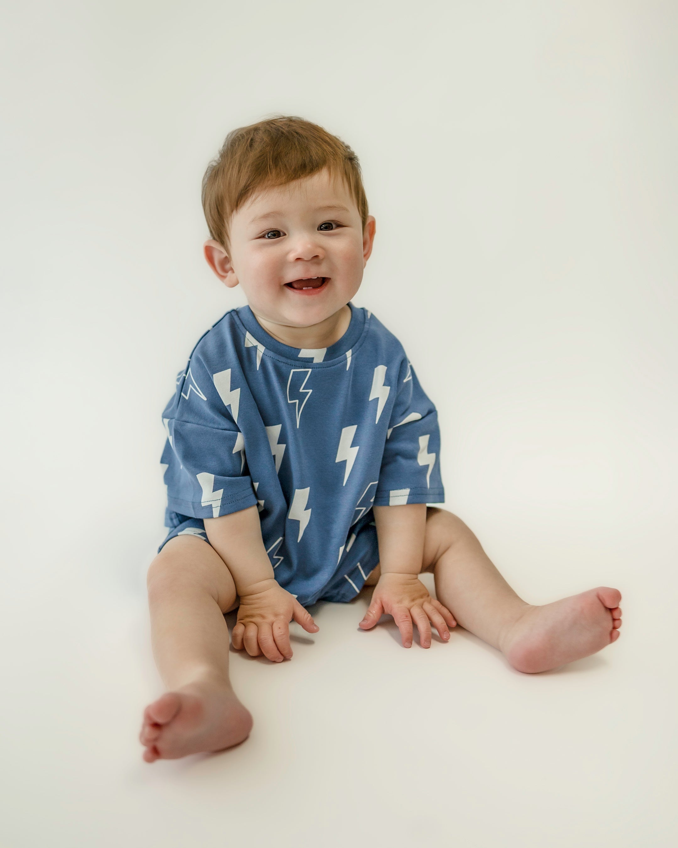 Smiling baby in oversized blue bolt print tee, sitting on a white background, showcasing playful summer style.