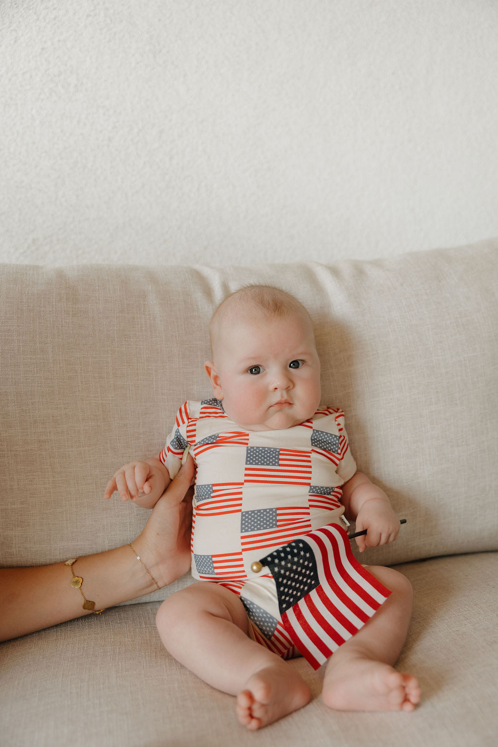 Baby in patriotic outfit holding mini American flag, sitting on beige couch, perfect for 4th of July celebrations.