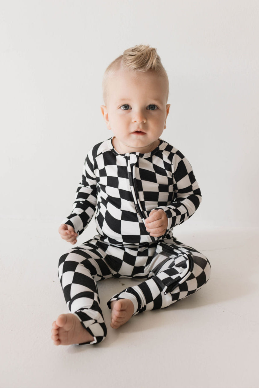 Baby wearing black and white wavy checkerboard bamboo zip pajamas, seated with a playful expression.