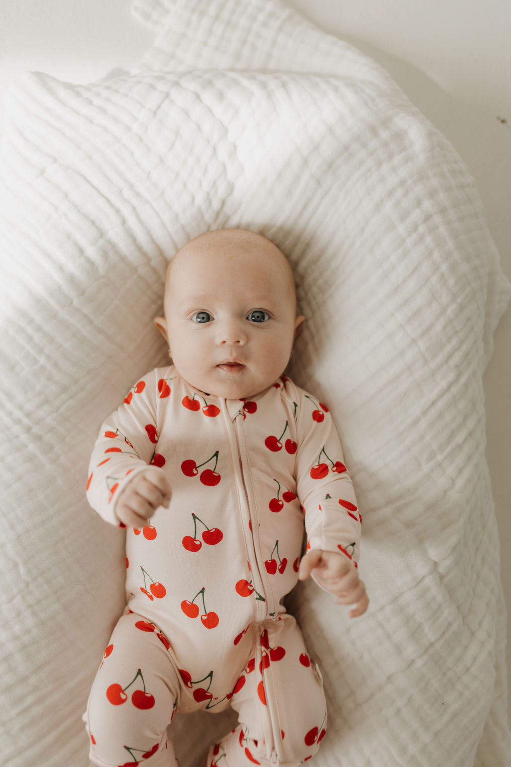 Infant wearing Cherry Orchard bamboo zip pajamas with a cherry print, laid on a soft blanket.