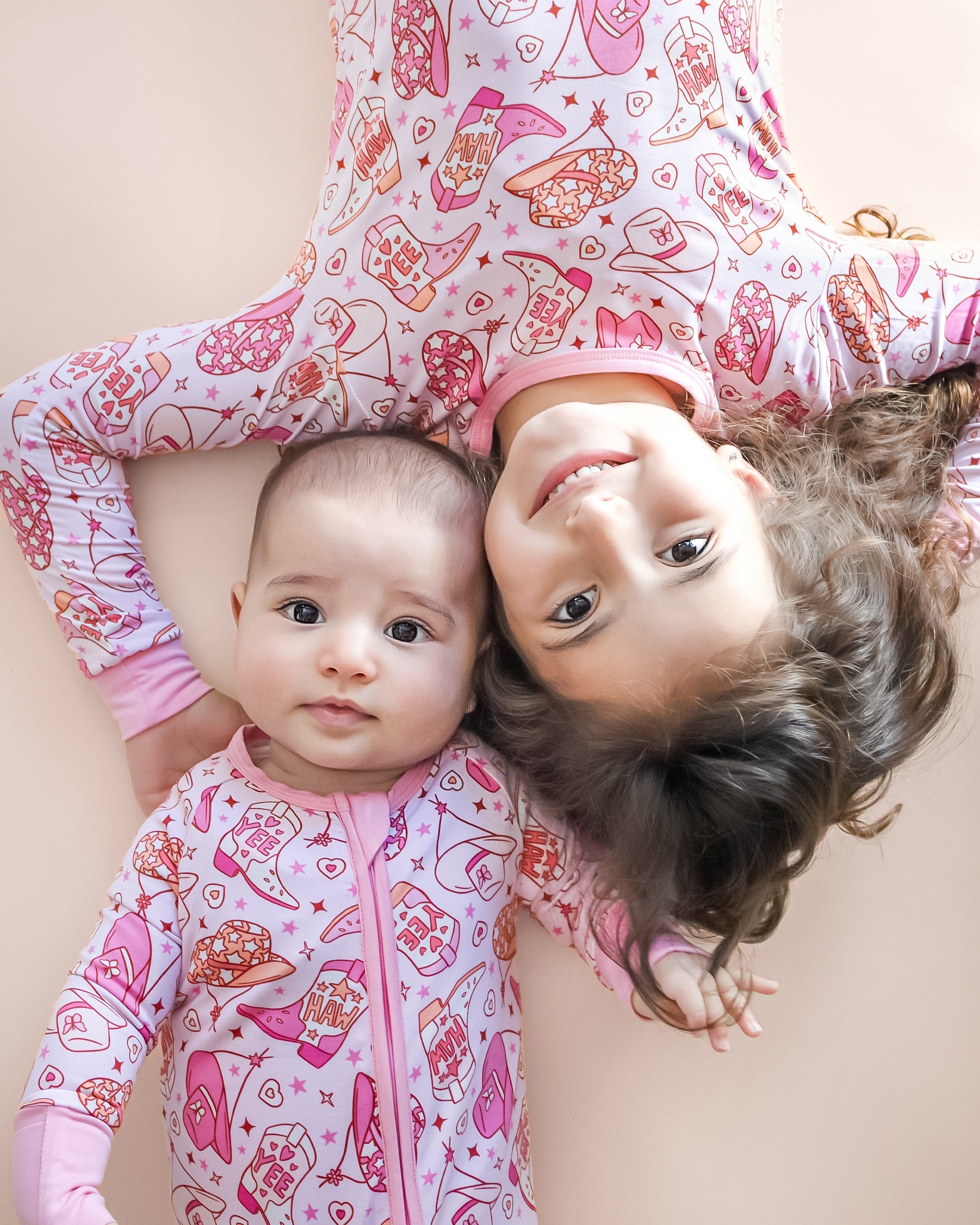 Baby and toddler wearing cowgirl bamboo zip rompers with playful western prints, smiling together on a soft background.