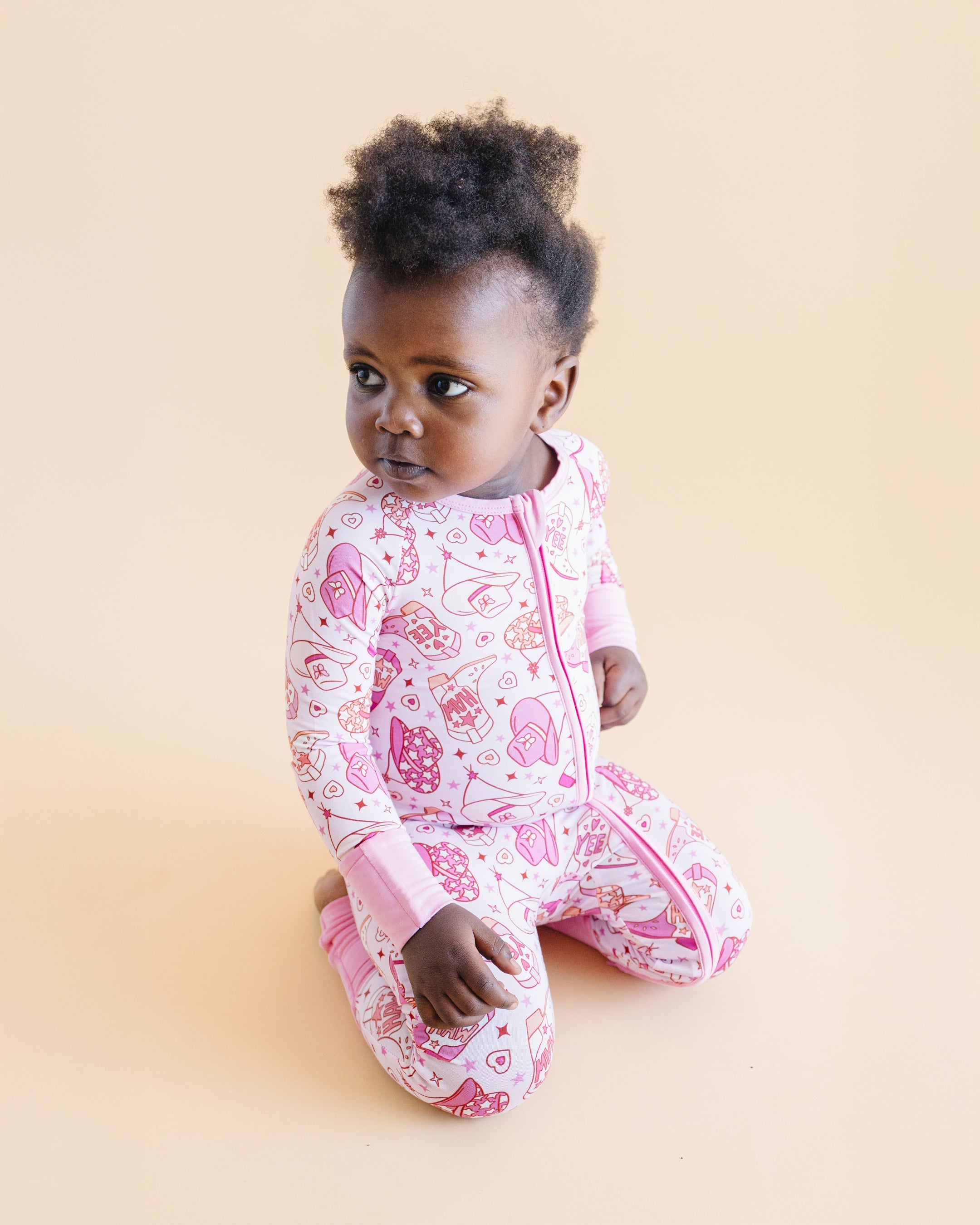 Baby wearing a pink cowgirl bamboo zip romper with playful western print, sitting on the floor.