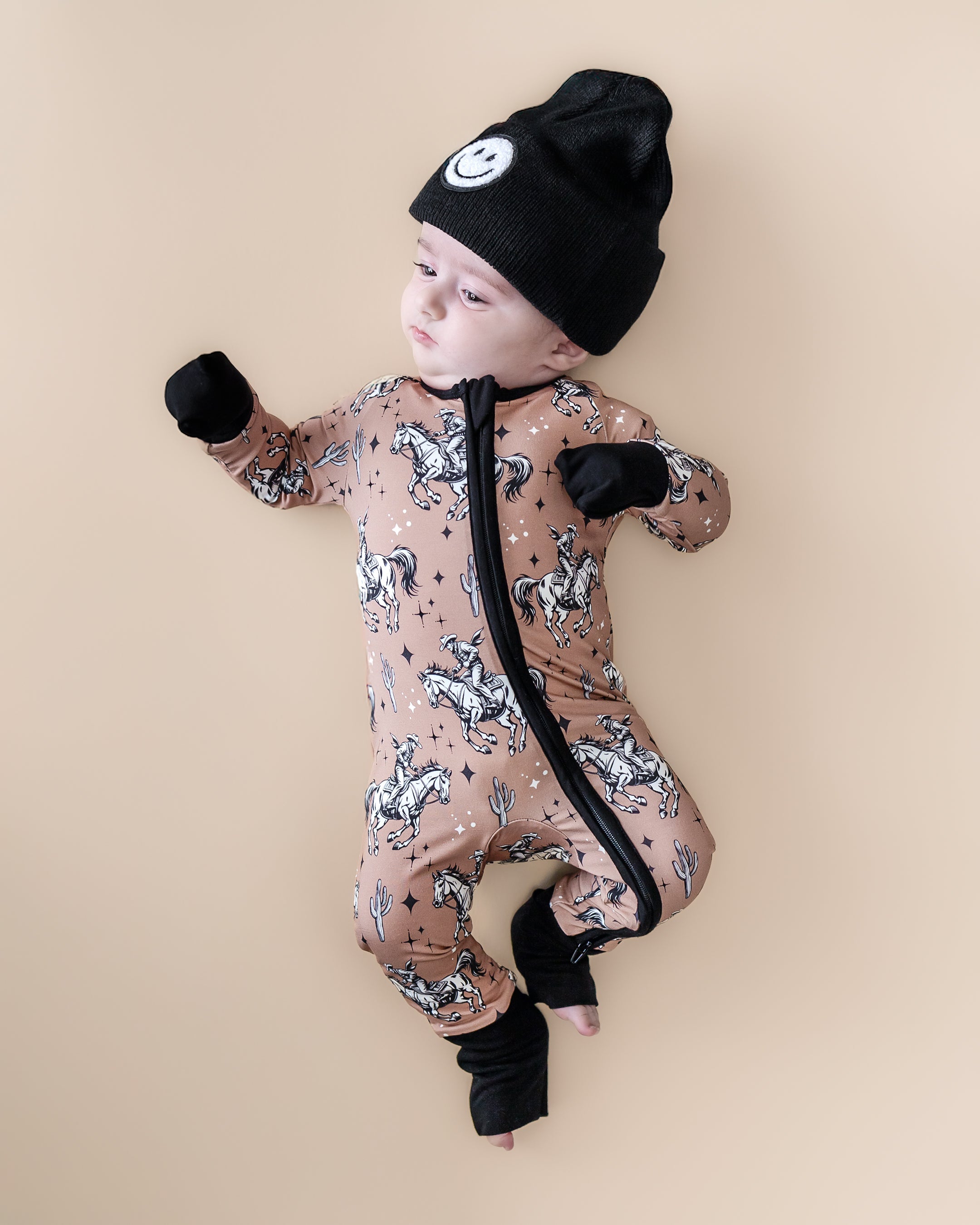 Baby wearing a Bamboo Zip Romper in cowboy print with a black beanie, on a sandy background.