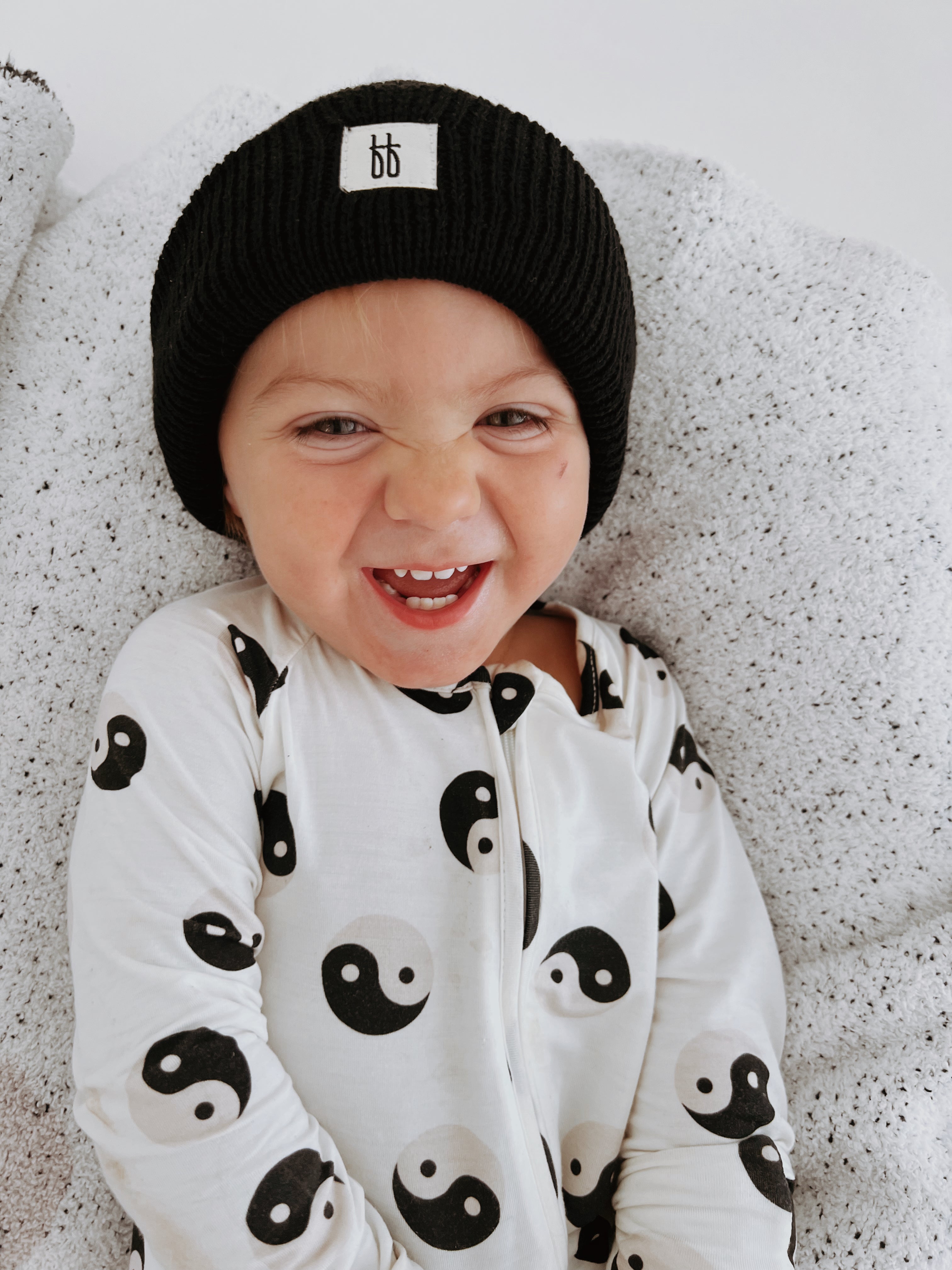 Happy baby wearing black & white yin yang bamboo zip pajamas with a black knit hat, snuggled on a cozy blanket.