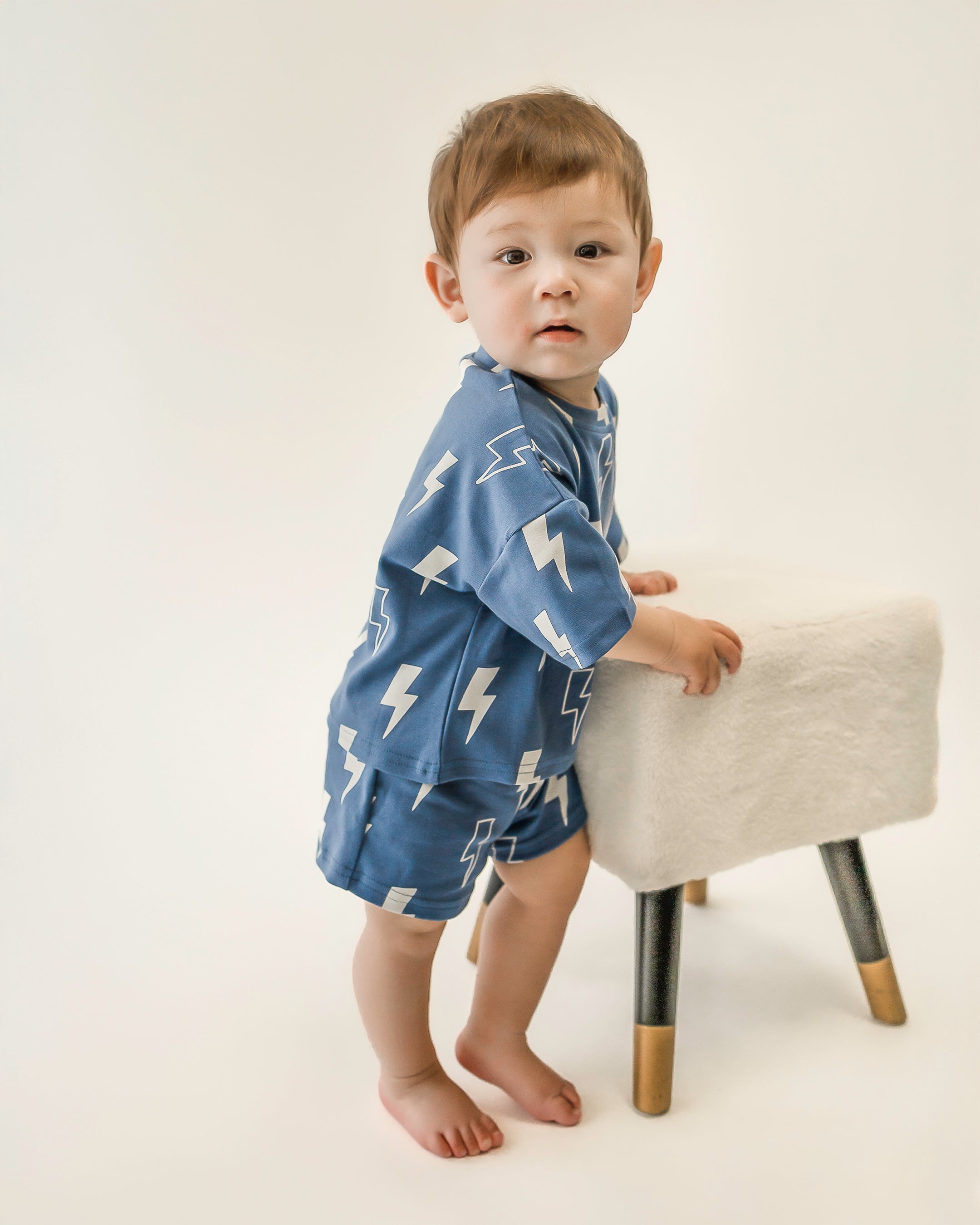 Toddler wearing blue bolt shorts set, featuring an oversized tee and matching shorts, standing beside a stool.