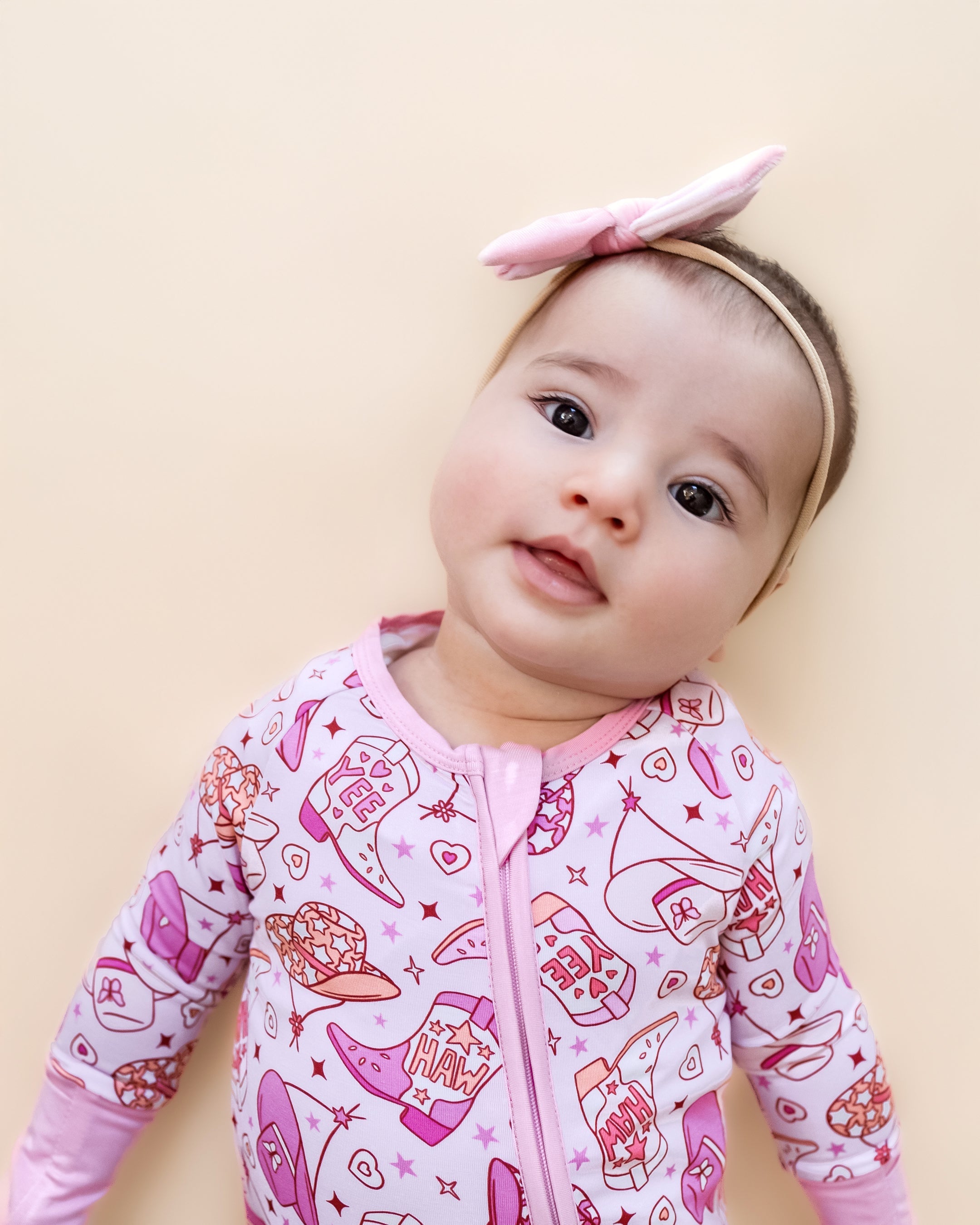 Baby wearing cowgirl bamboo zip romper with playful print and headband, striking a cute pose.