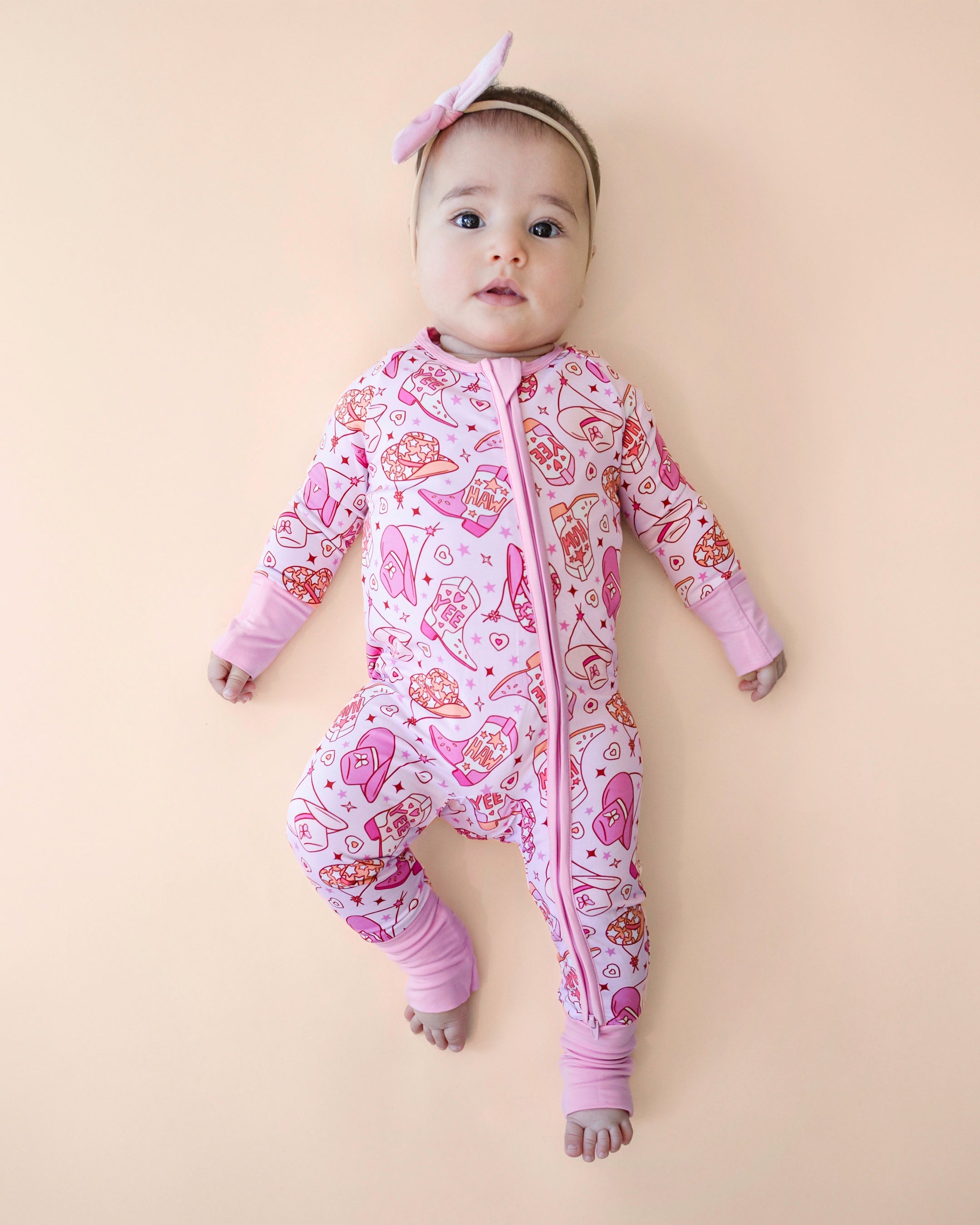 Baby in cowgirl bamboo zip romper with playful western print, featuring boots and hats, on a light peach background.
