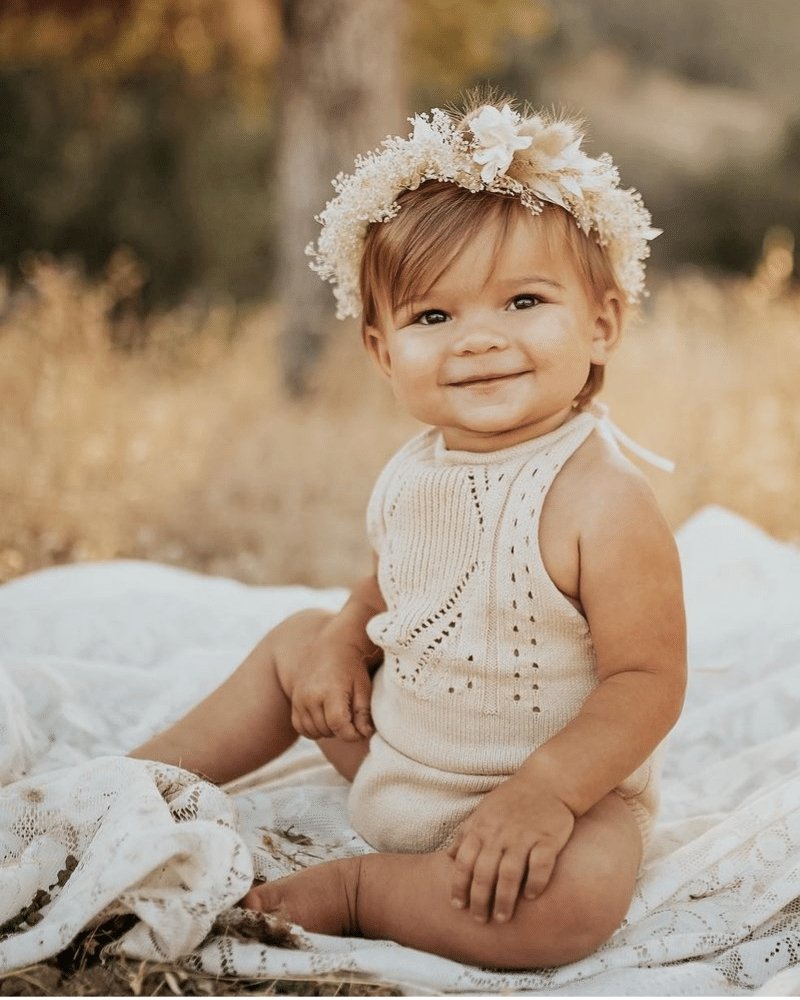 Adorable baby in Drake crochet halter romper, smiling while surrounded by natural scenery and wearing a floral crown.