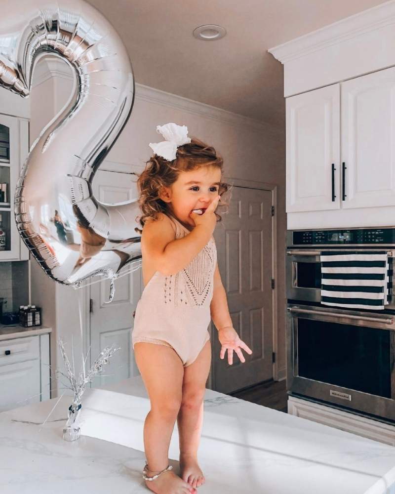 Toddler in a crochet romper celebrating 2nd birthday with silver balloon on a kitchen counter.