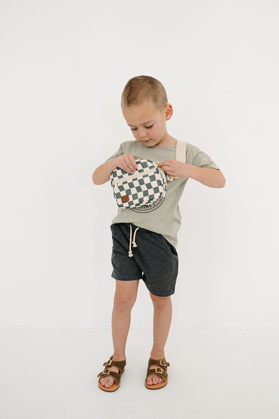 Child wearing gray shorts and shirt, holding a green and white checkered mini fanny pack, fashion for kids.