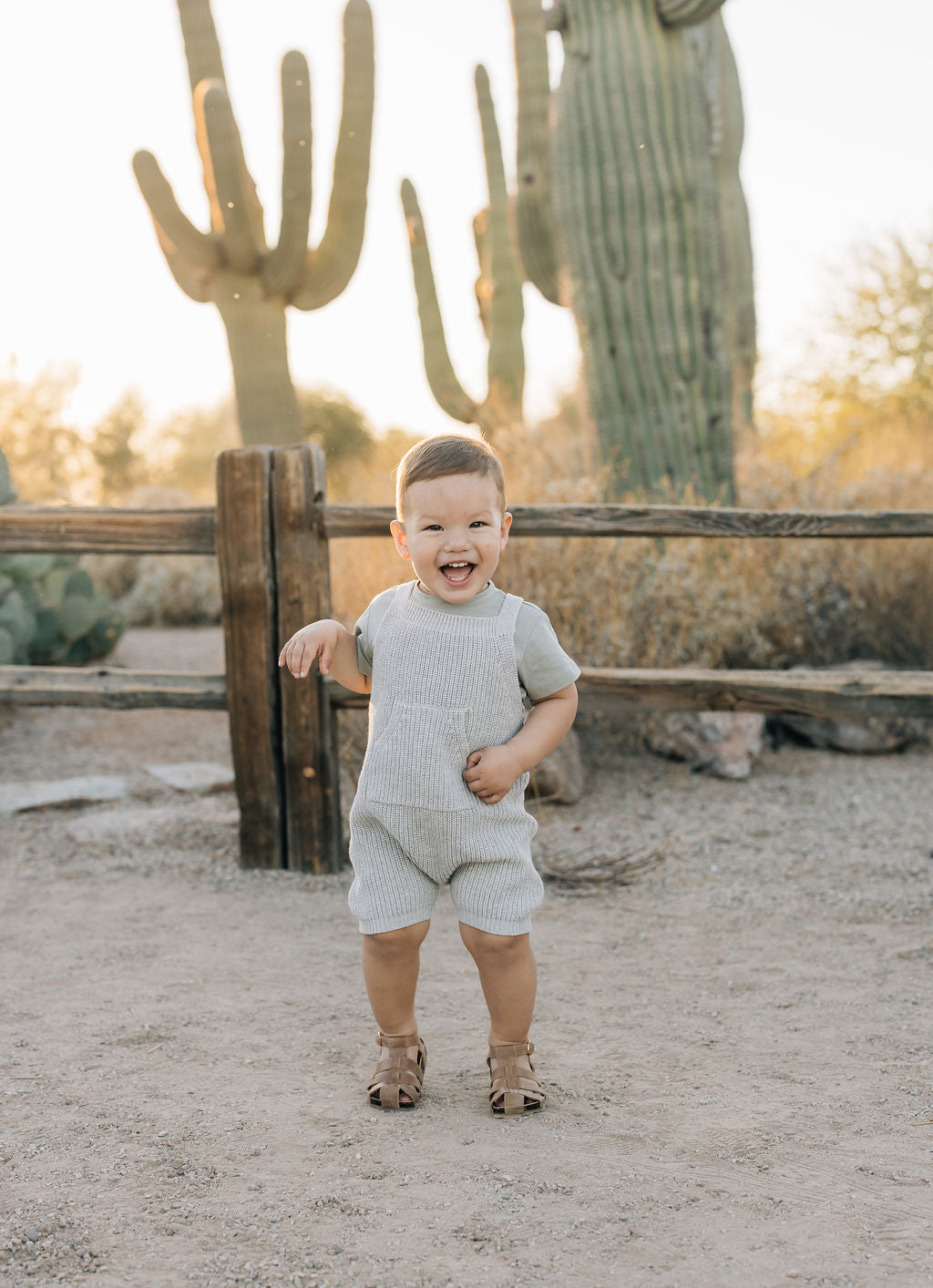 Happy baby wearing heather grey knit overalls, playing outdoors in a desert setting with cacti.