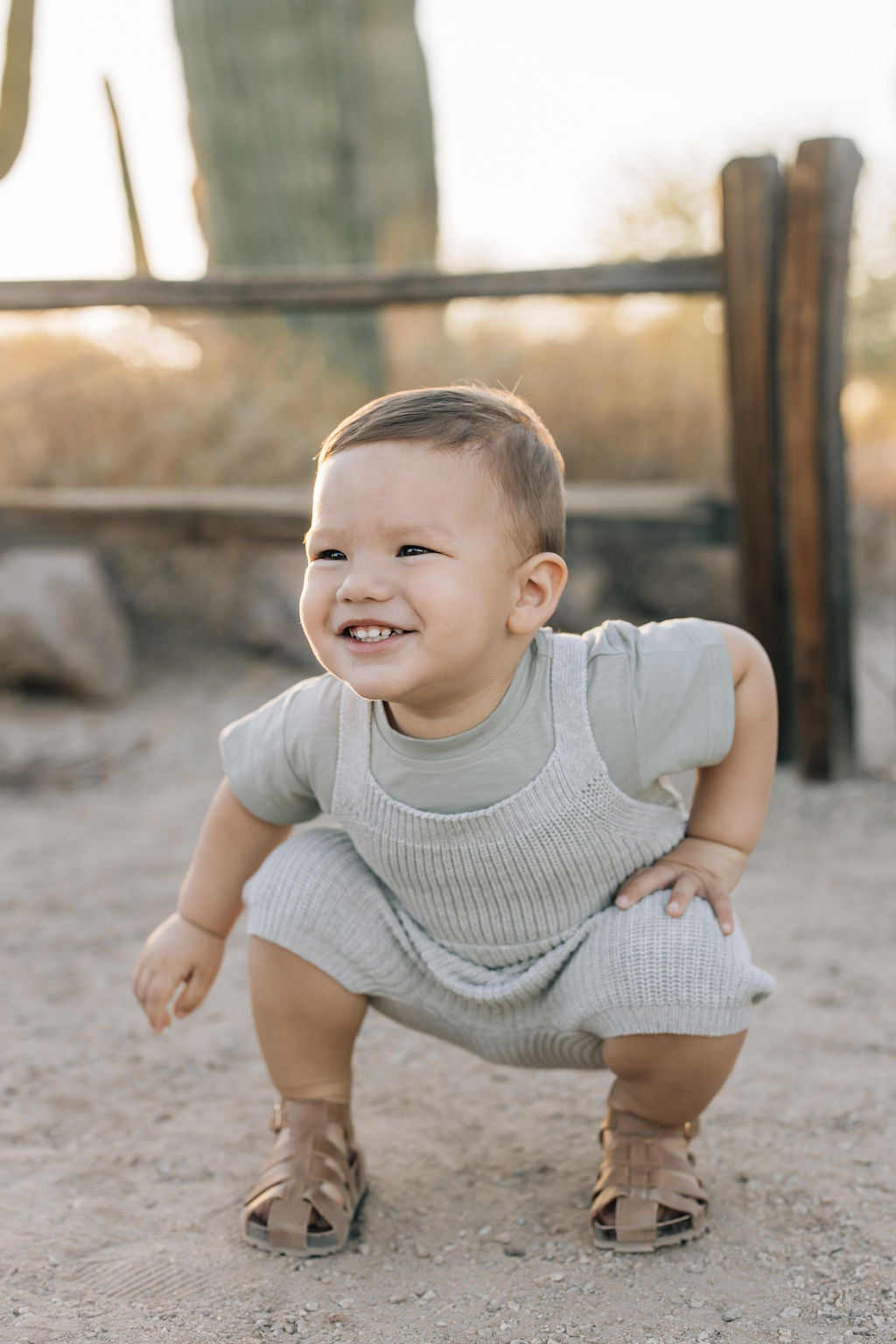 Baby wearing heather grey pocket knit overalls, smiling while squatting outdoors.