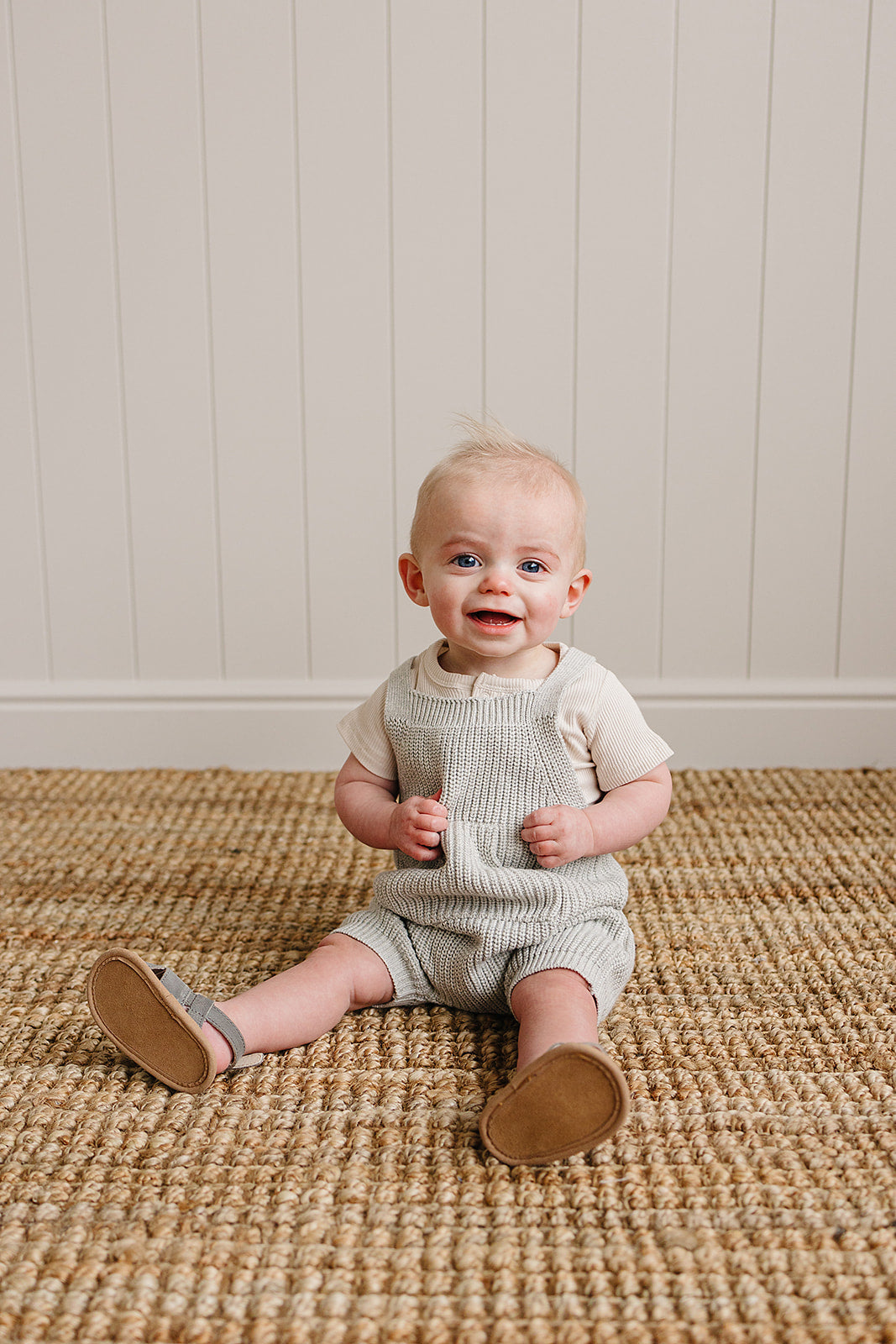 Adorable baby wearing heather grey pocket knit overalls, seated on a textured rug, smiling happily.