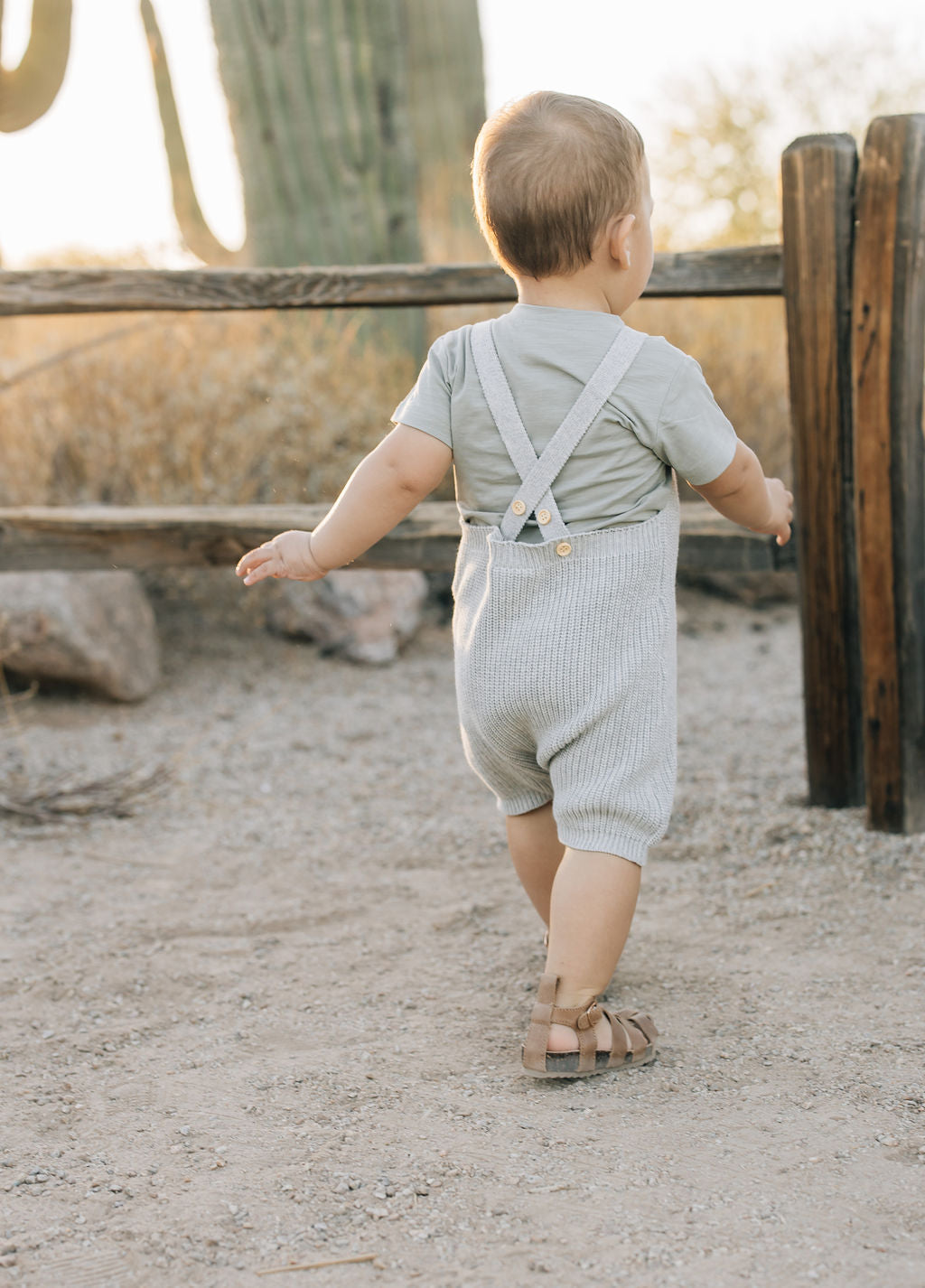 Toddler wearing heather grey knit overalls with button straps, playing outdoors near a wooden fence.