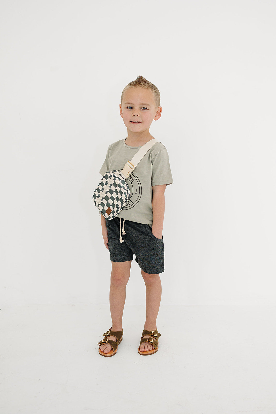 Boy wearing a green checkered mini fanny pack with a t-shirt and shorts, in a bright setting.