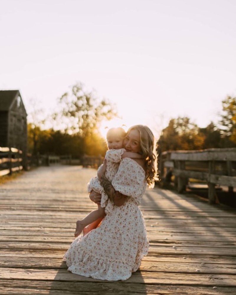 Mother and child wearing matching yellow floral dresses on a sunny wooden path.