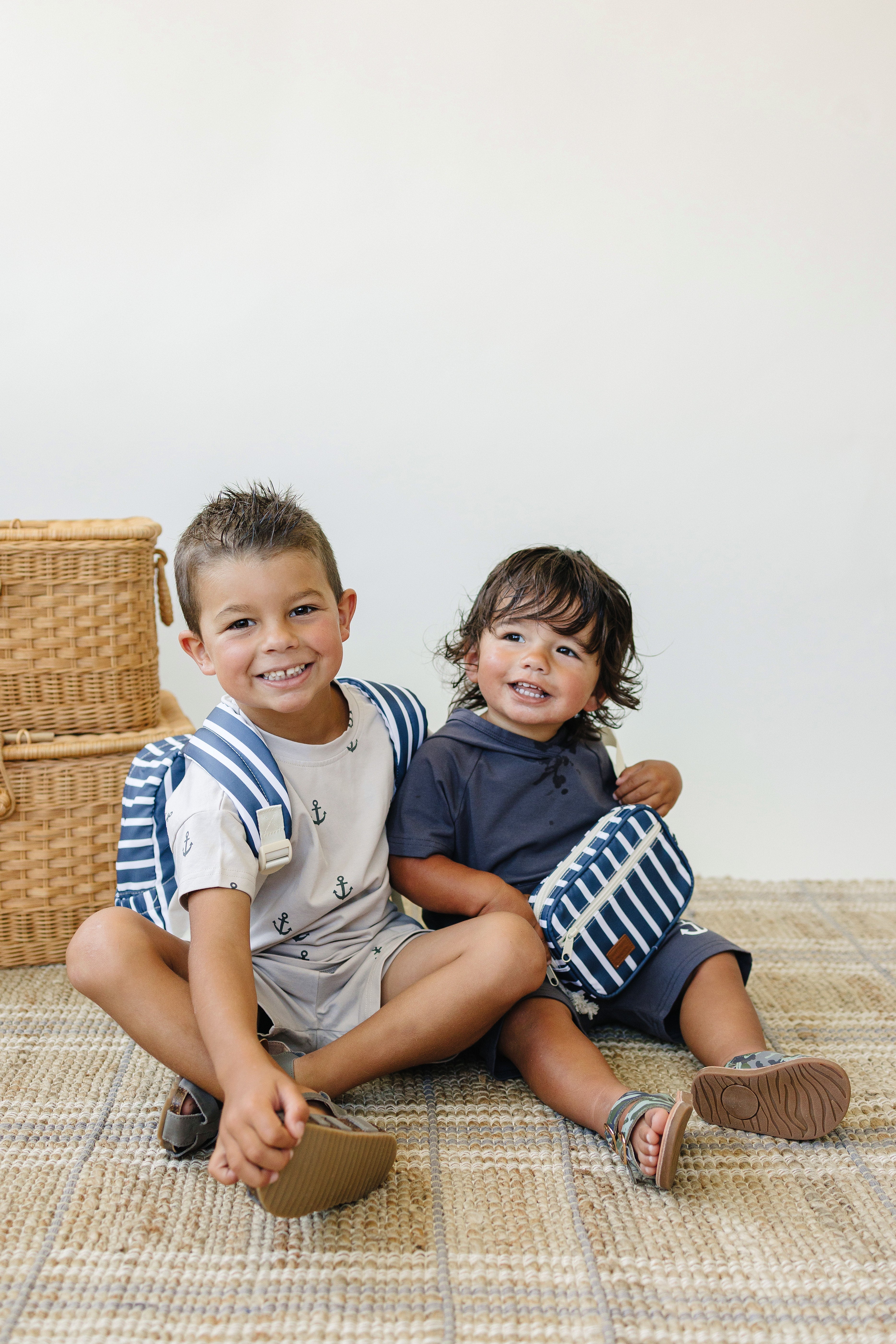 Two kids sitting on a rug, wearing navy and white striped mini fanny packs and cheerful outfits.