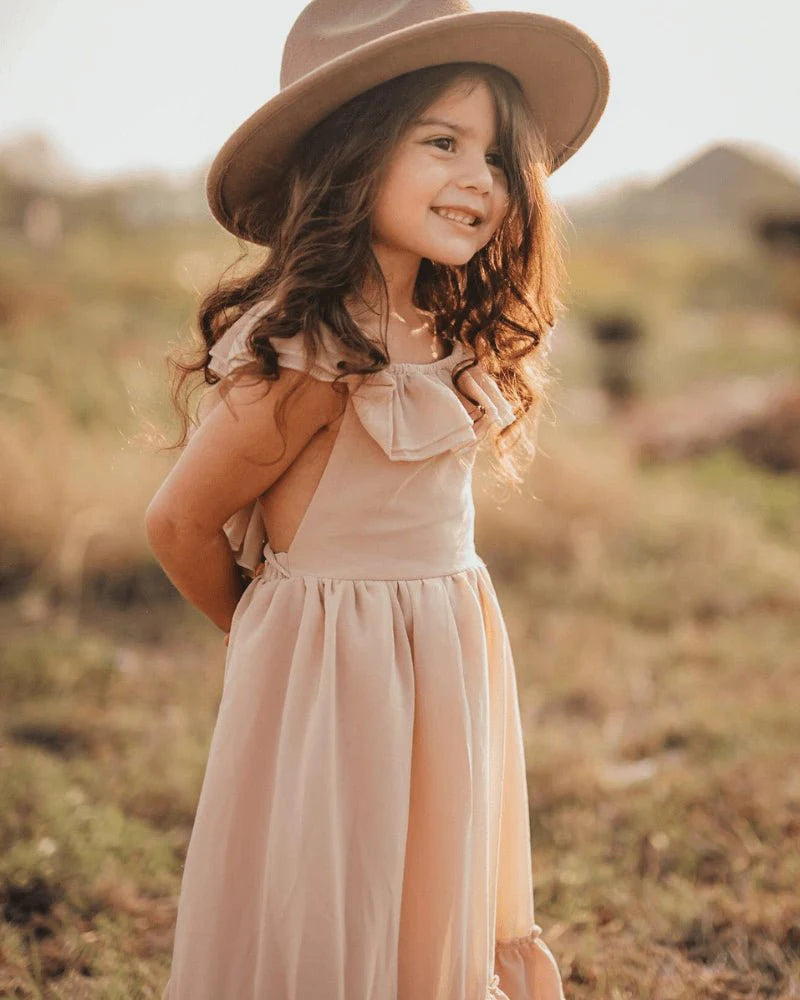 A young girl wearing a champagne dress with layered ruffles and a hat, smiling in an outdoor setting.