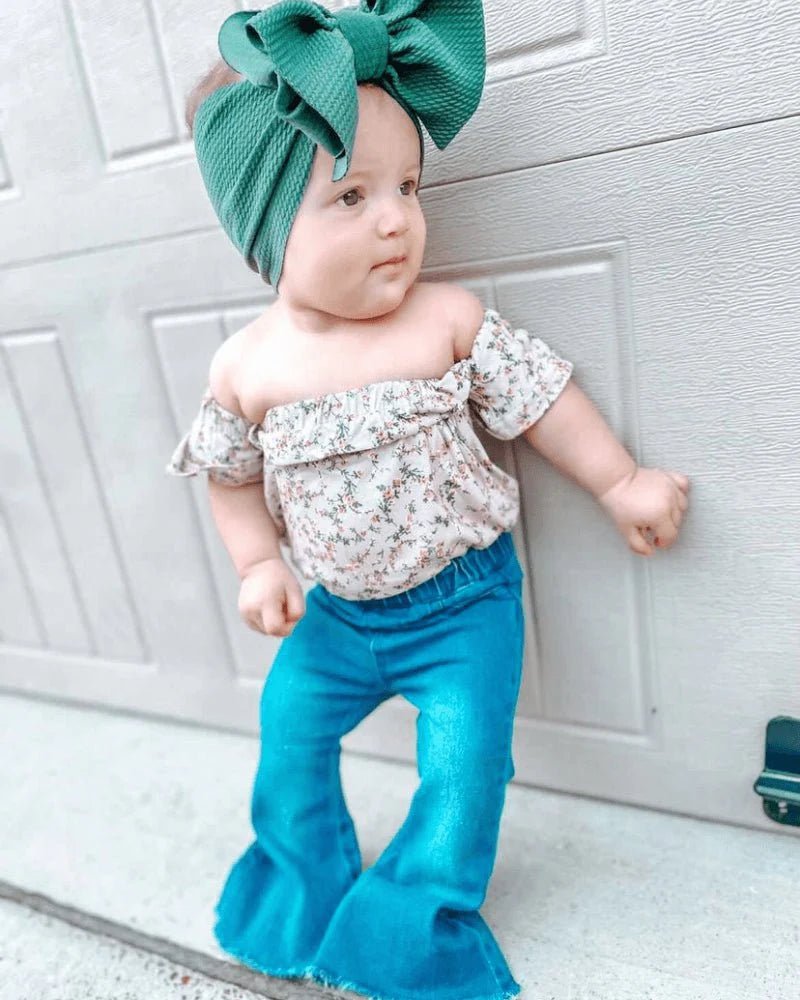 Toddler wearing floral off-shoulder top and teal bell bottom jeans with a green headband, posing against a garage door.