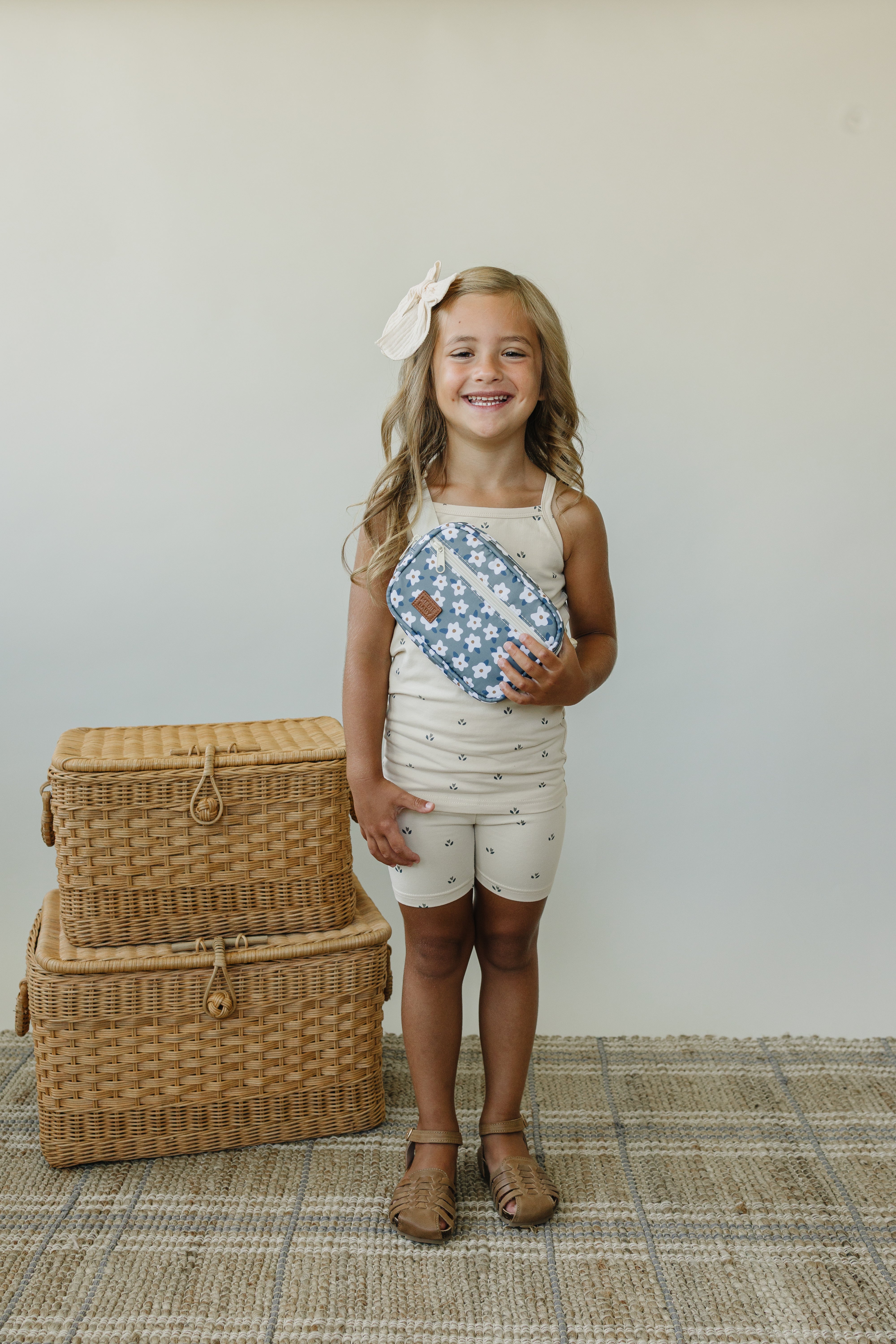 Smiling girl holding a blue floral mini fanny pack, wearing a light outfit, with baskets in the background.
