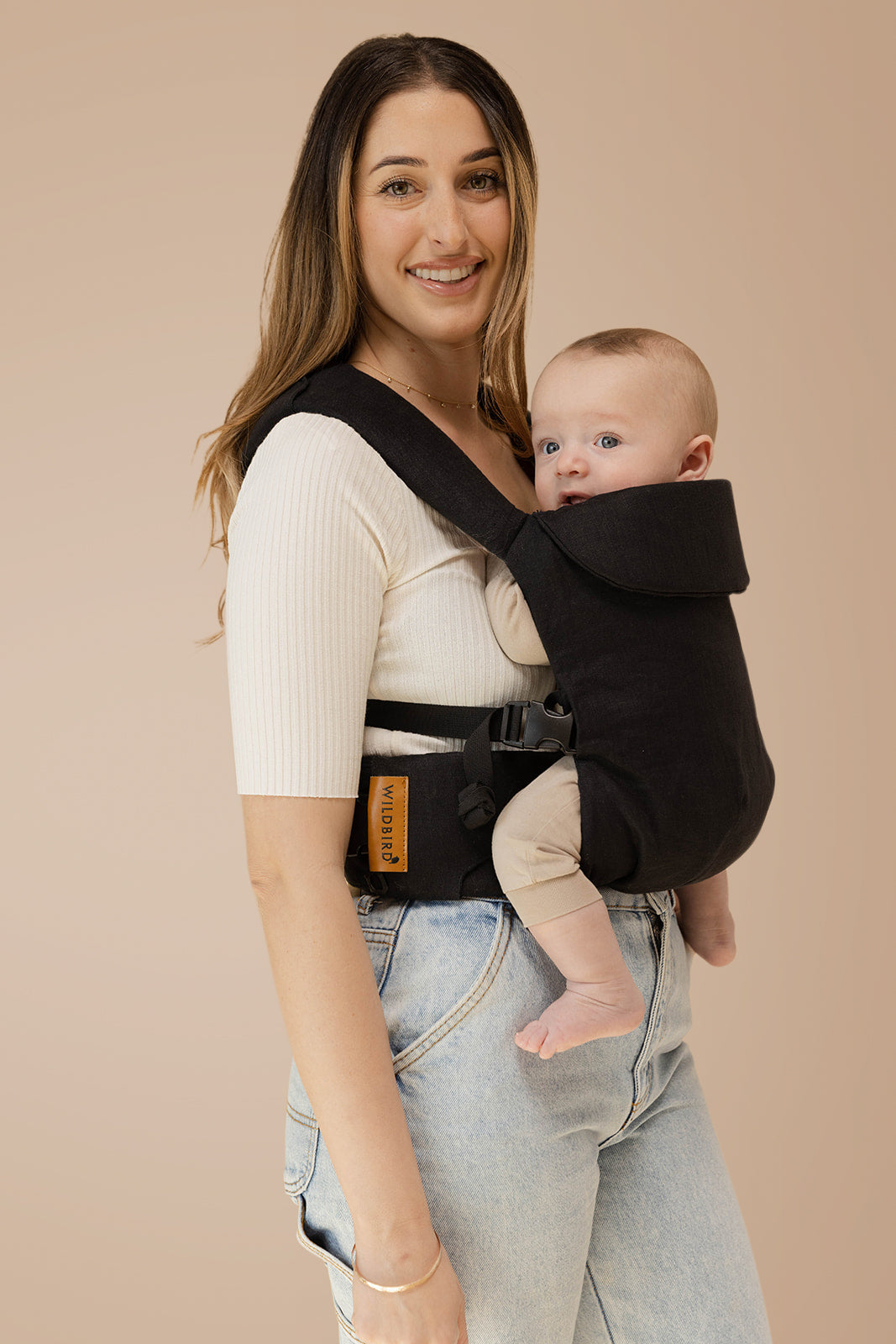 Woman wearing Raven Aerial Carrier, a sleek black baby carrier, comfortably holding a baby, showcasing its modern design.