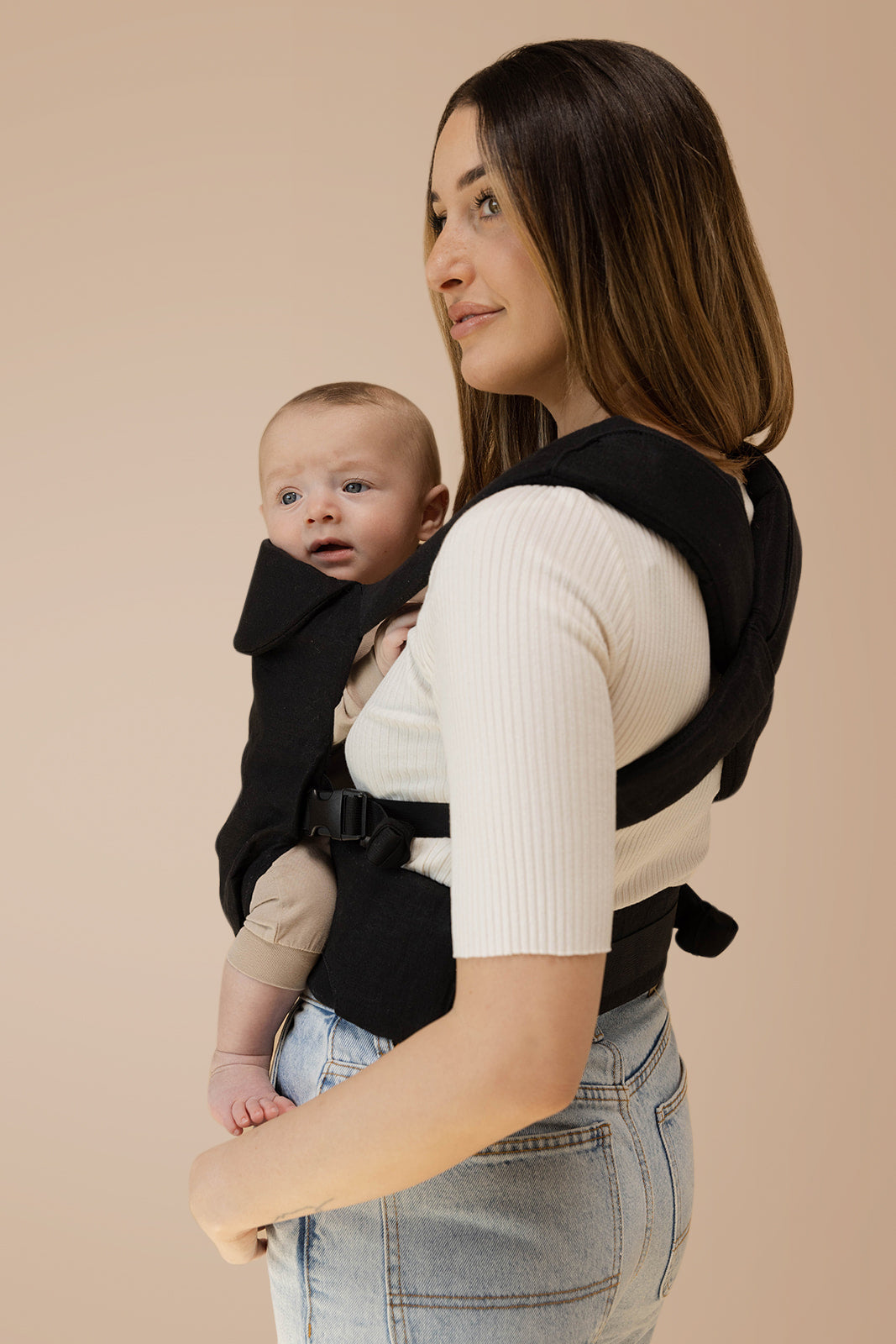 A mother wearing the Raven Aerial Carrier, holding her baby with a sleek monochrome design, perfect for caregivers.