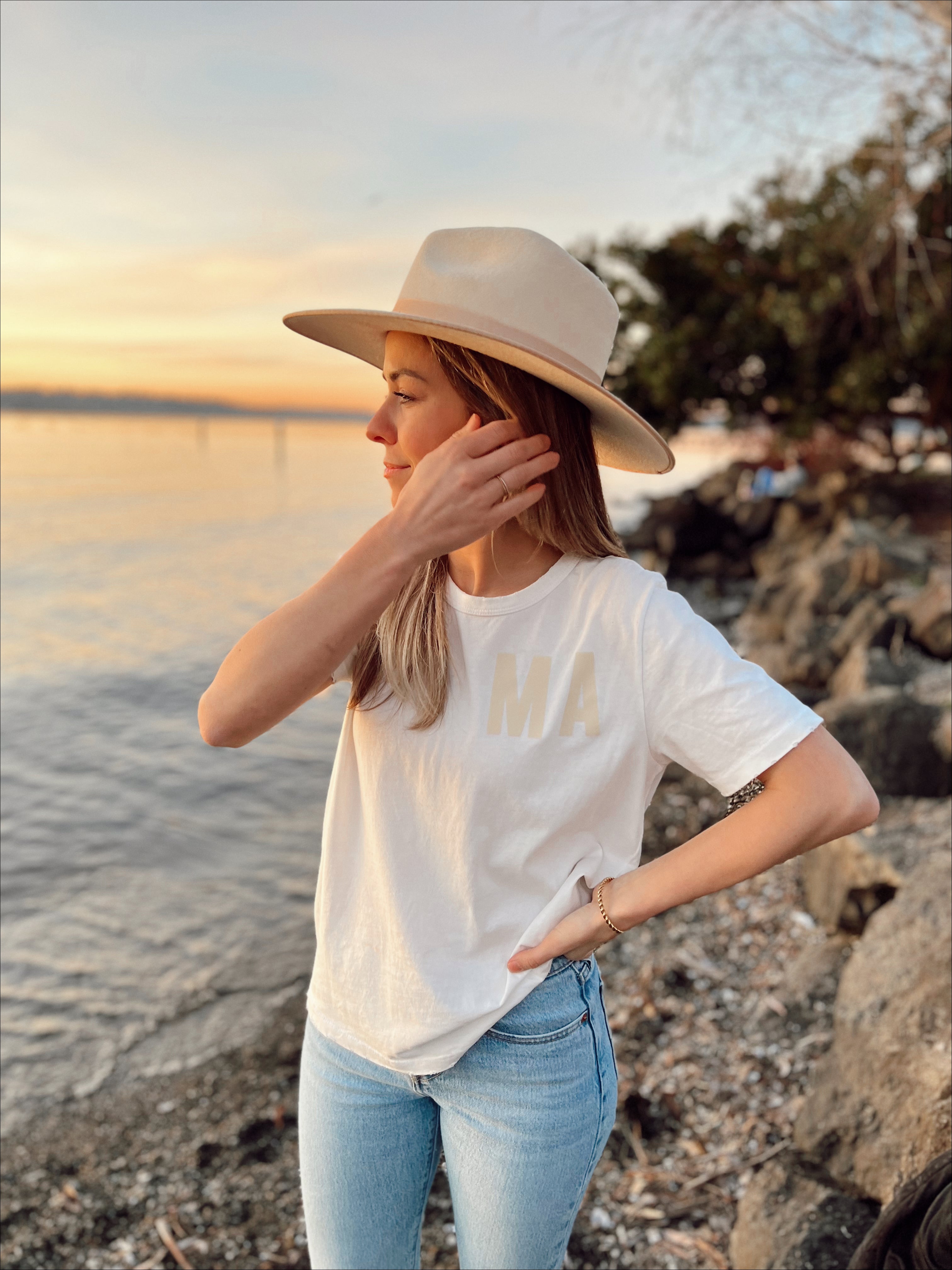 Woman wearing Organic Vintage MA Tee and wide-brim hat by the riverside at sunset, showcasing relaxed style.