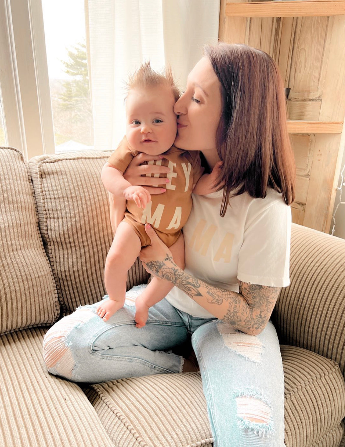 Mom and baby on a couch, wearing matching 'Hey Mama' outfits, showcasing family bonding and comfort.