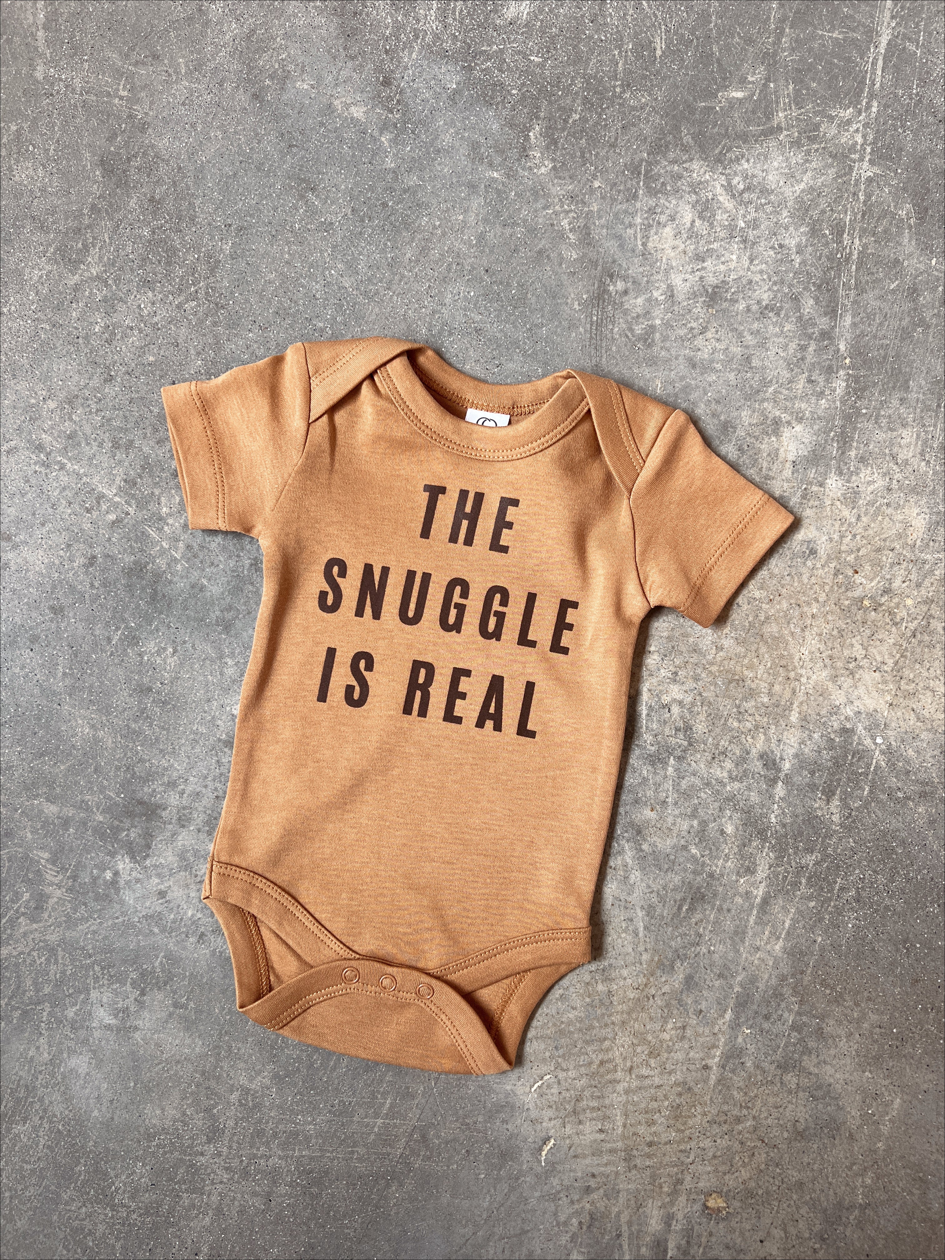 The Snuggle Is Real organic baby onesie in soft brown fabric, featuring minimalist black text on a cozy bodysuit.
