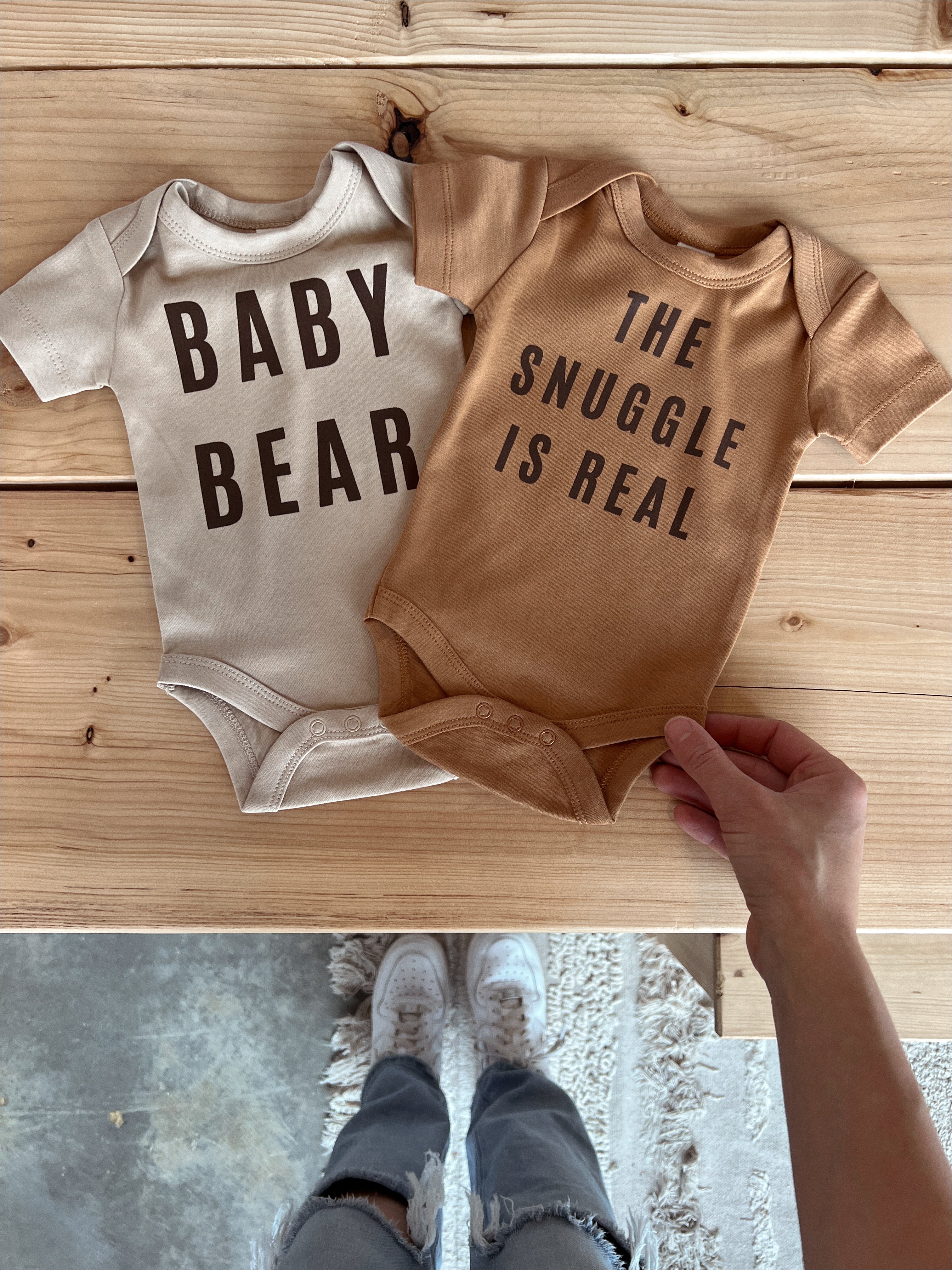 Two organic baby onesies with prints: 'Baby Bear' and 'The Snuggle Is Real', showcased on a wooden table.
