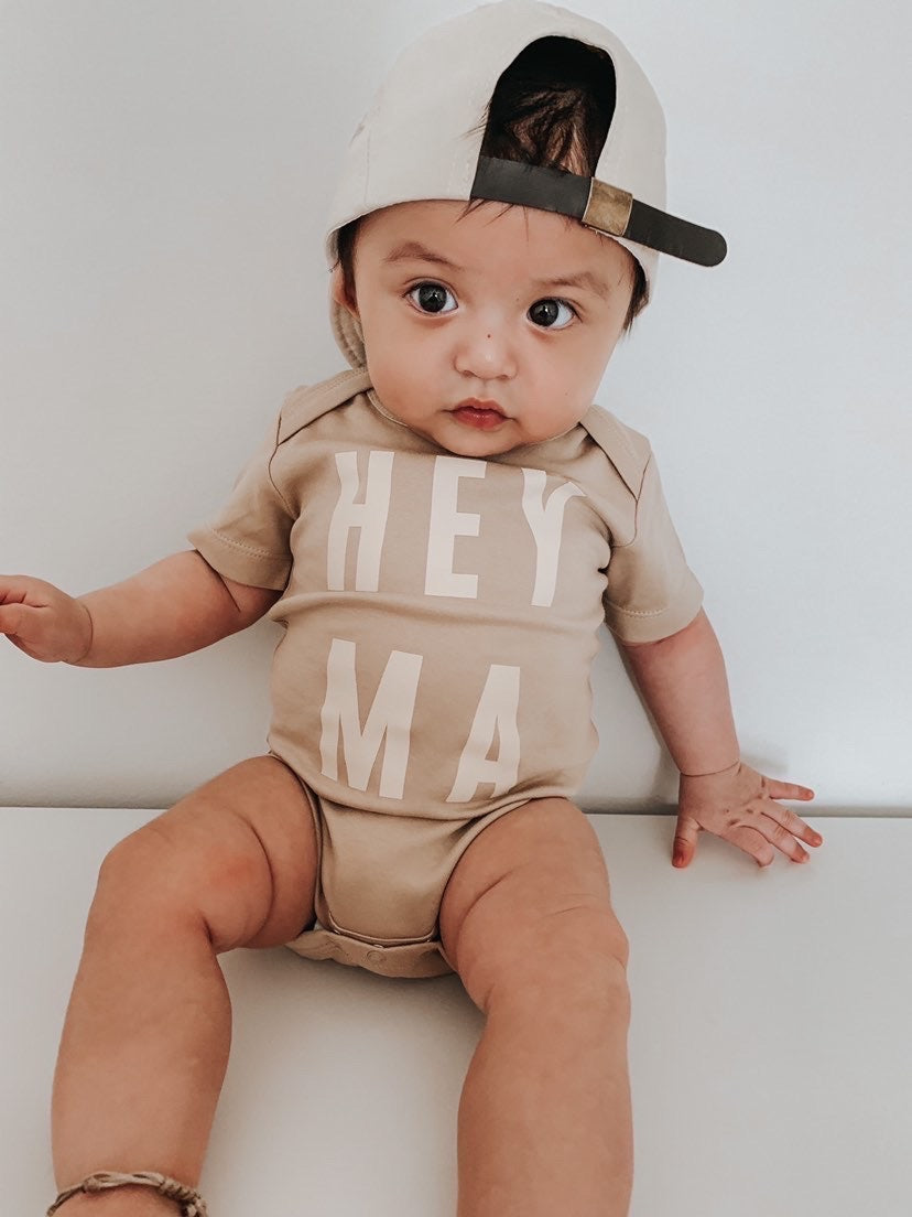 Baby in cream organic onesie with 'Hey Ma' design and cap, showcasing trendy and cool baby style.