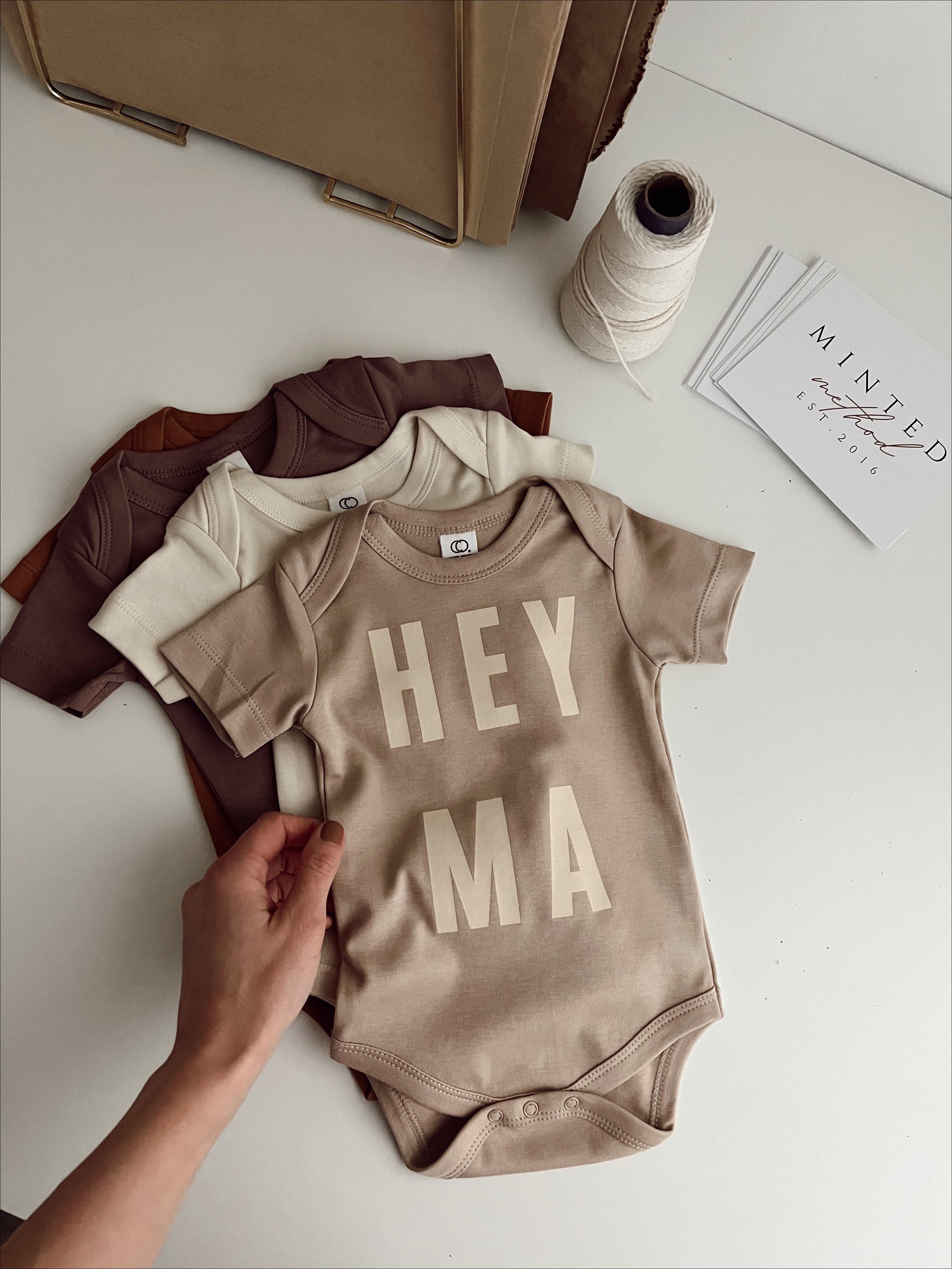 Cream organic baby onesies with "Hey Ma" design, showcasing trendy vintage style for cool moms and little ones.